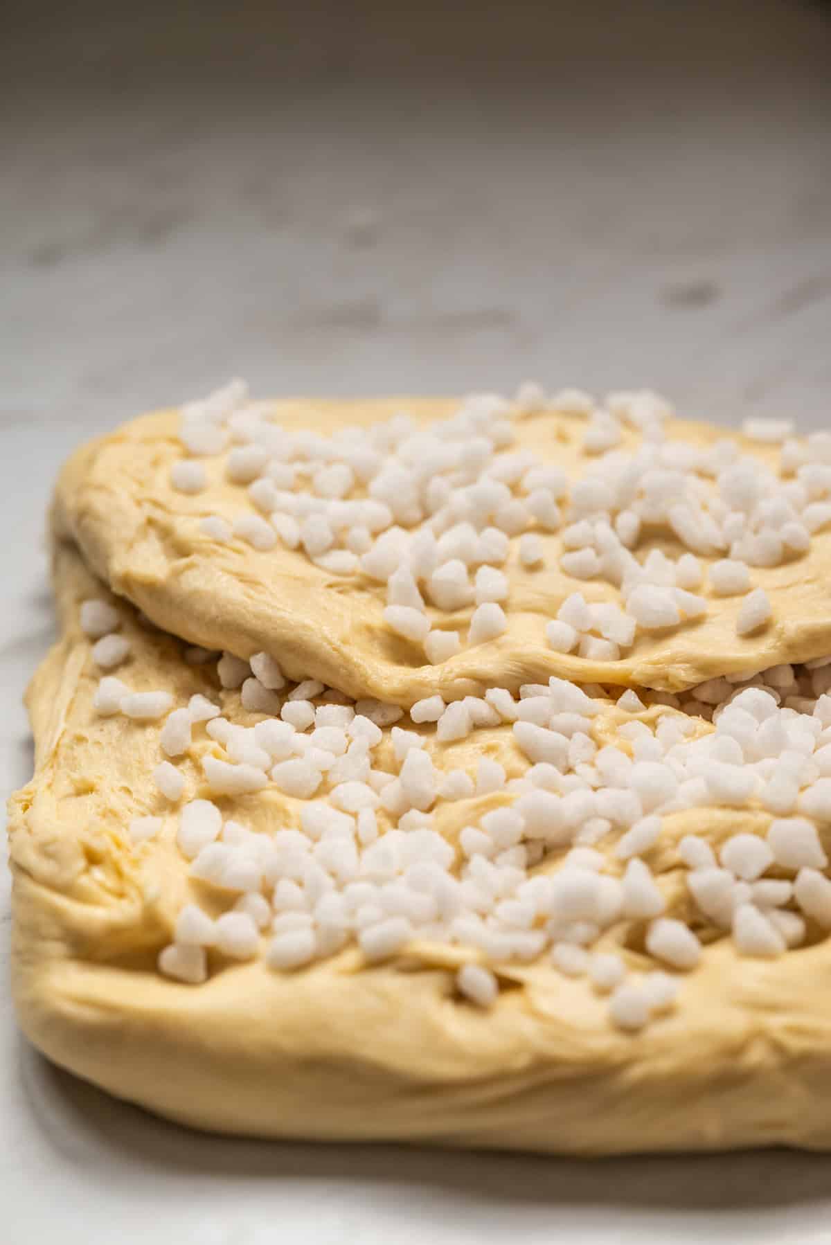 A close-up of sweet bread dough topped with pearl sugar, resting on a marble surface, ready to be baked.