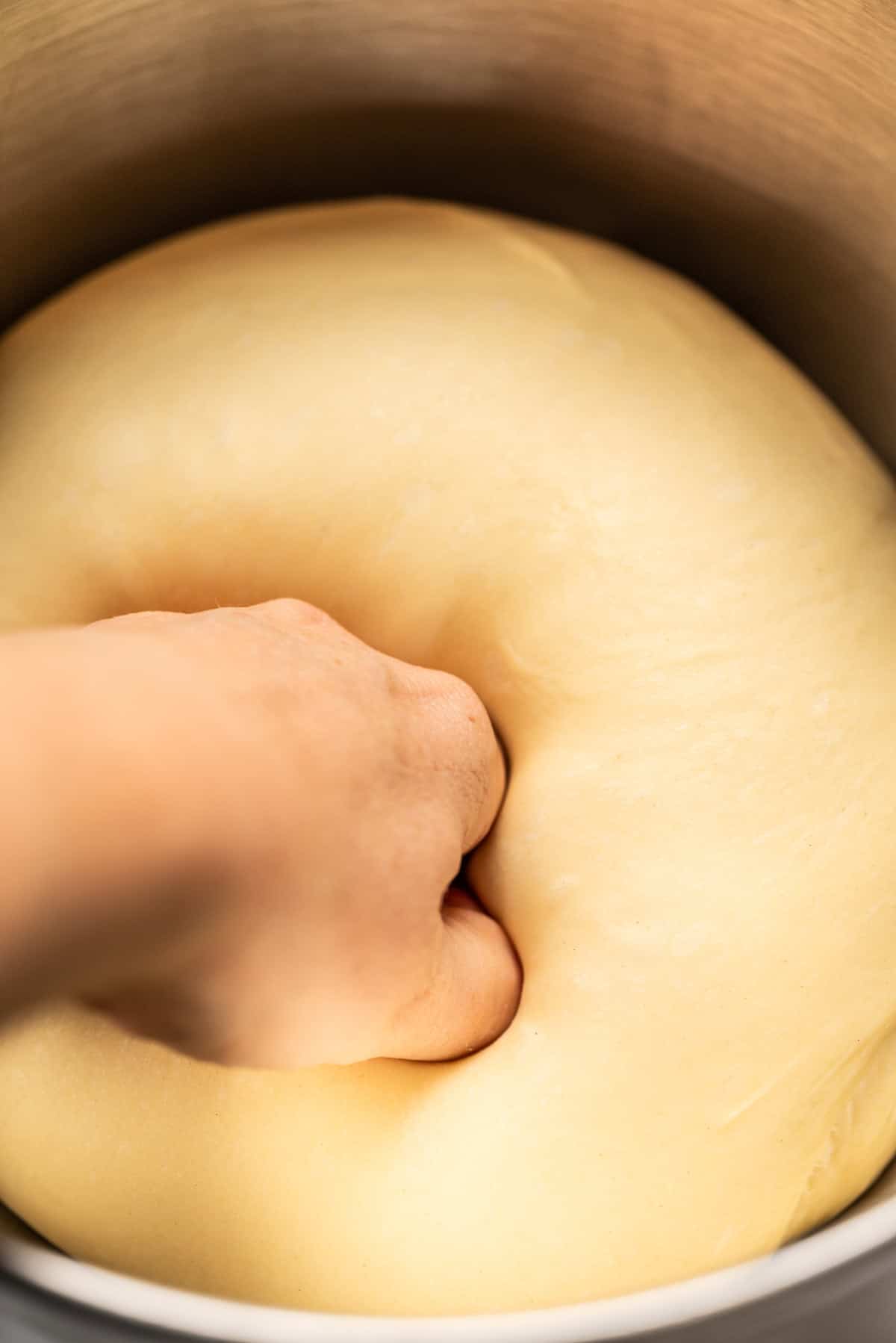 A hand presses into a ball of risen dough inside a metal bowl, showing the dough being punched down after rising.
