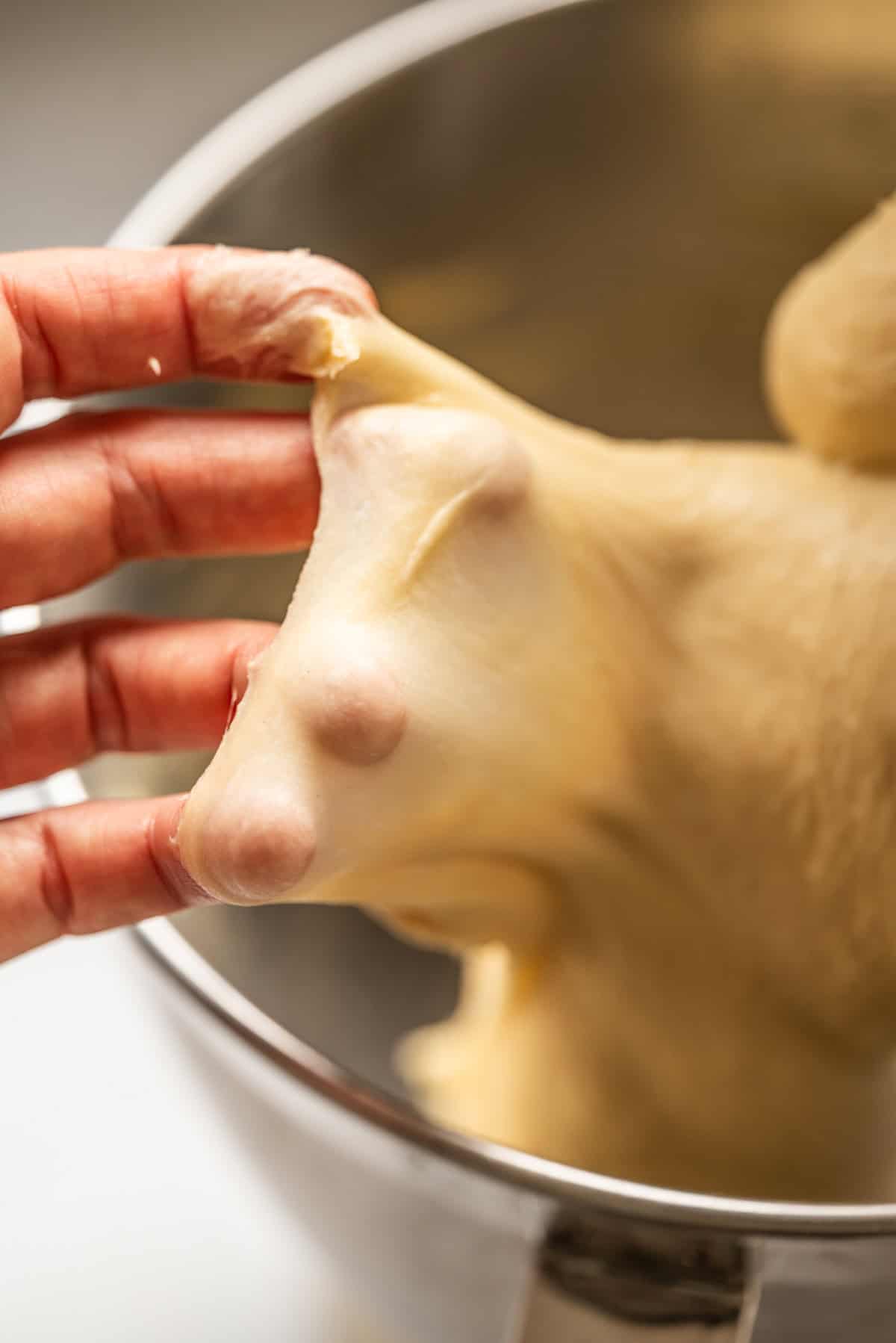 A hand stretches a piece of smooth, elastic bread dough above a metal mixing bowl, showing the dough’s thin, translucent texture.