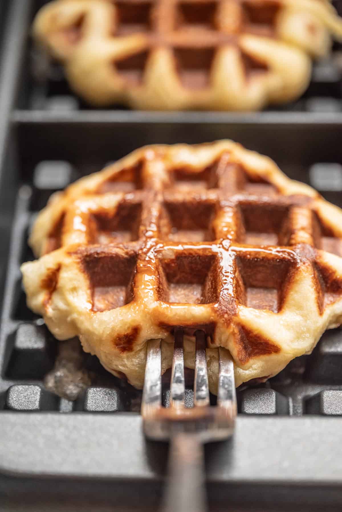A close-up of a golden-brown waffle on a waffle iron, with a metal fork lifting it. Another waffle is visible in the background. The waffle appears fresh and slightly shiny from melted butter or syrup.