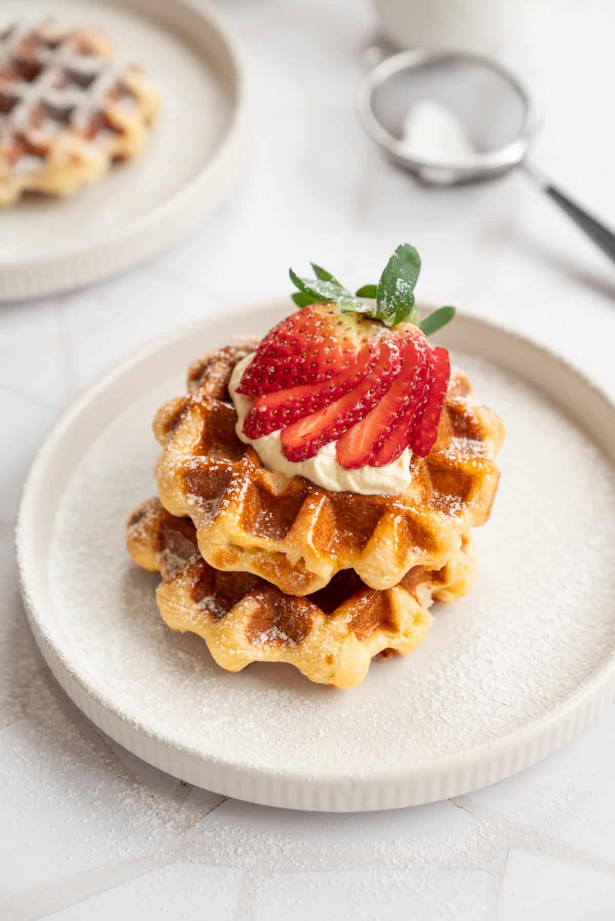 A stack of two Liège waffles topped with whipped cream and sliced strawberries sits on a white plate dusted with powdered sugar. A metal sifter and another plate with waffles are blurred in the background.