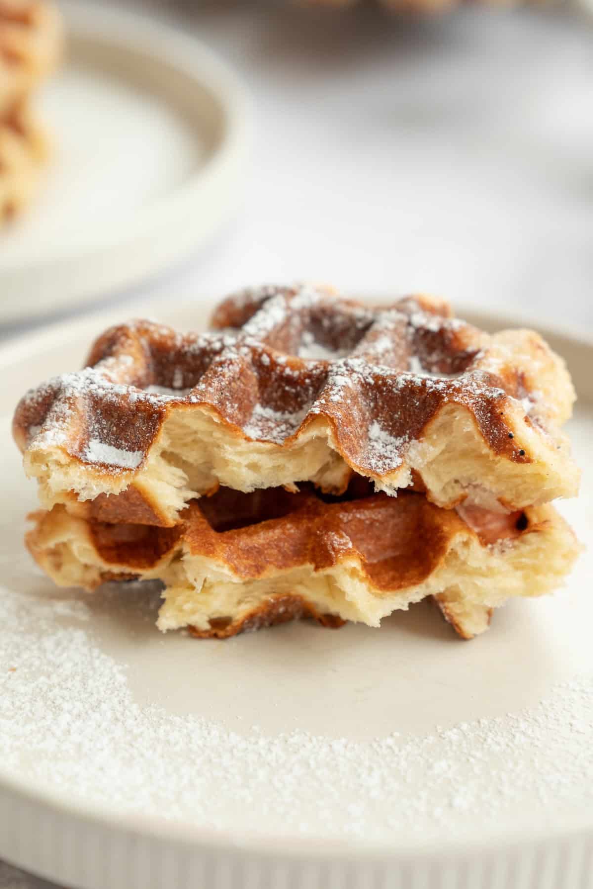 Two stacked pieces of golden, crispy Liège waffles dusted with powdered sugar sit on a light-colored plate.
