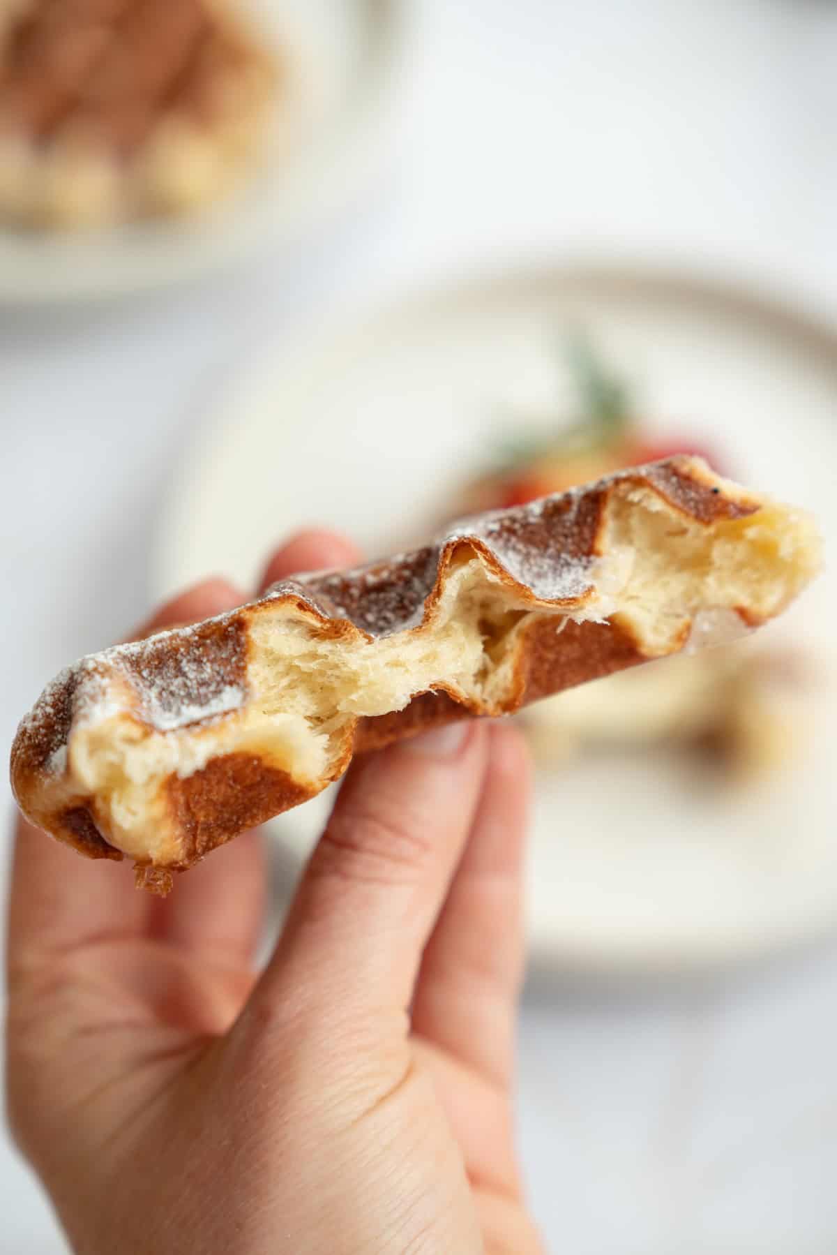 A hand holds up half of a torn, airy Liège waffle dusted with powdered sugar, showing its soft, fluffy interior. A blurred plate is visible in the background.