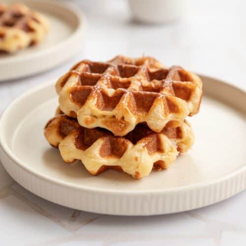 Two golden brown Liège waffles are stacked on a beige plate, placed on a light-colored table. Another plate with more waffles and a white mug are blurred in the background.
