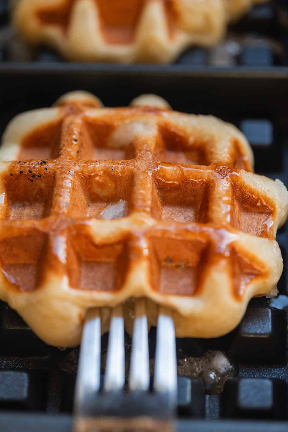 A close-up of a golden-brown Liège waffle being lifted from a waffle iron with a metal fork.