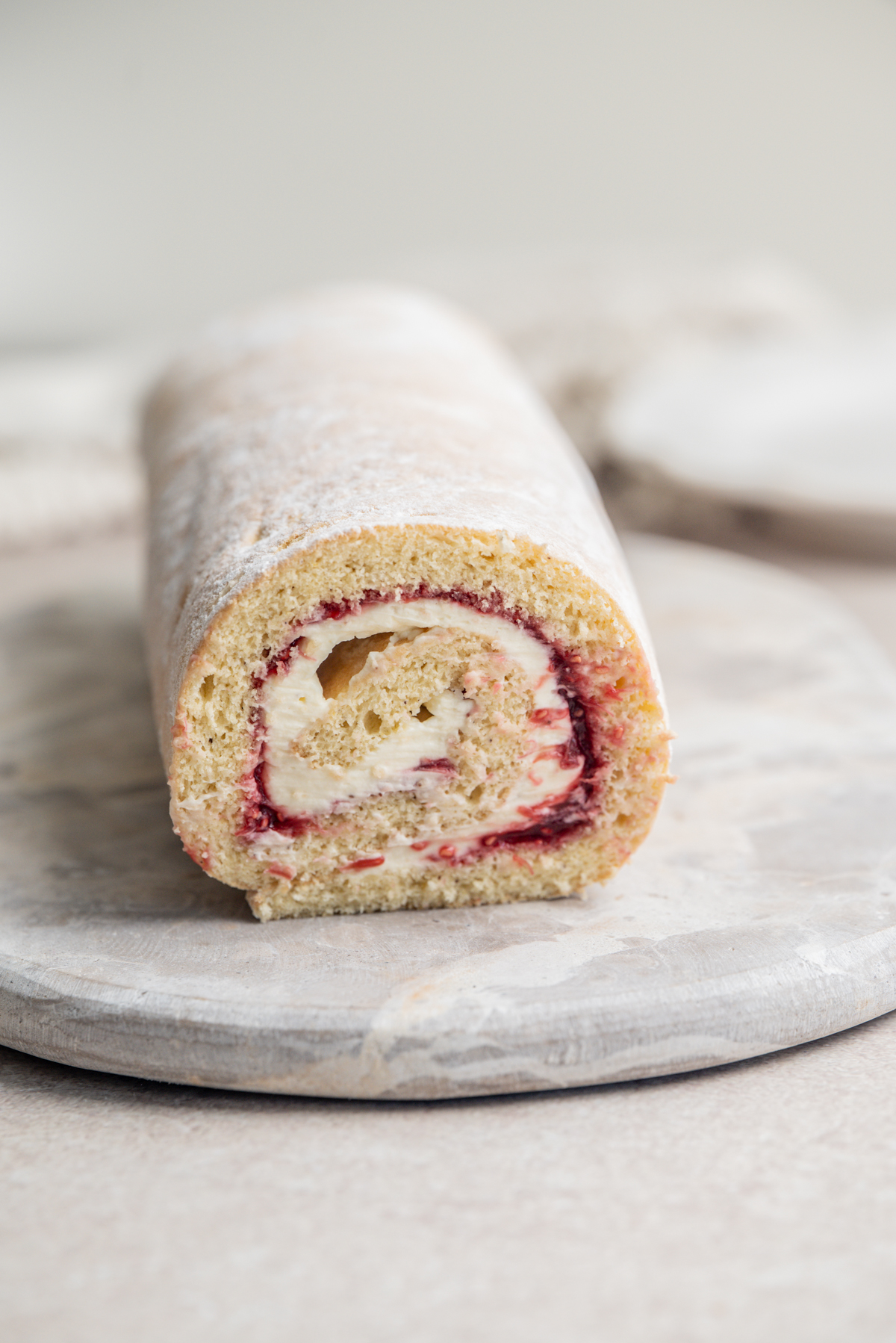 A Swiss roll cake with visible layers of yellow sponge, white cream, and red jam, sits on a round marble board.