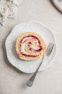 A slice of rhubarb raspberry Swiss roll cake with cream and berry jam filling on a white decorative plate, served with an ornate fork on a light-colored surface.
