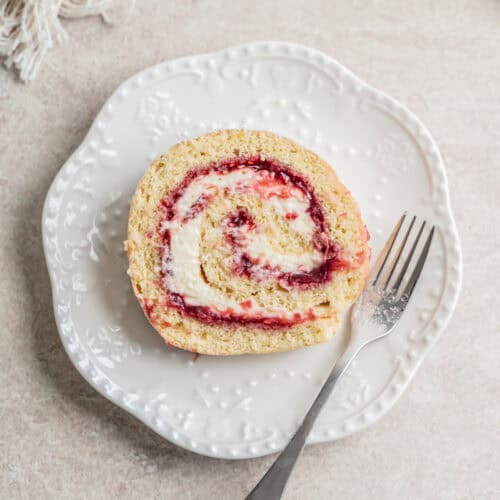 A slice of rhubarb raspberry Swiss roll cake with cream and berry jam filling on a white decorative plate, served with an ornate fork on a light-colored surface.