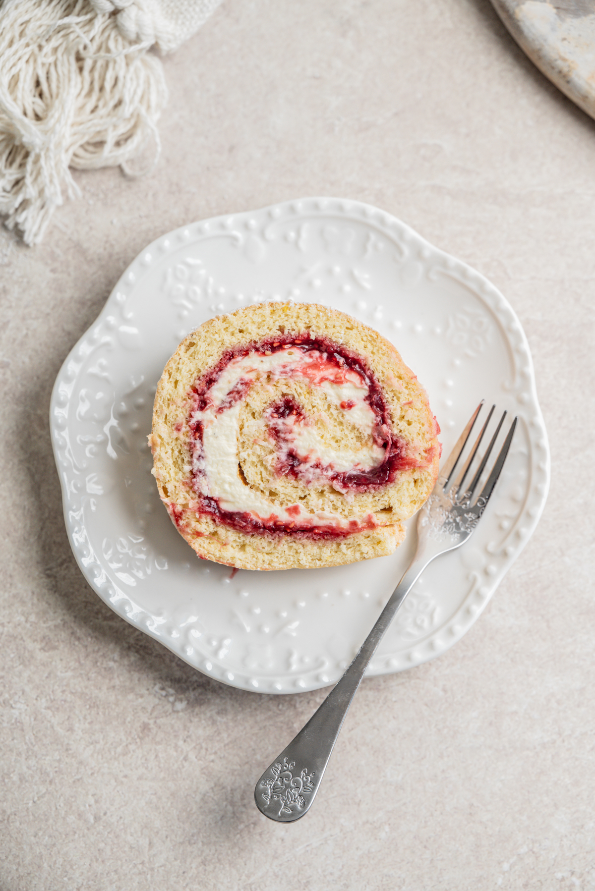 A slice of rhubarb raspberry Swiss roll cake with cream and berry jam filling on a white decorative plate, served with an ornate fork on a light-colored surface.