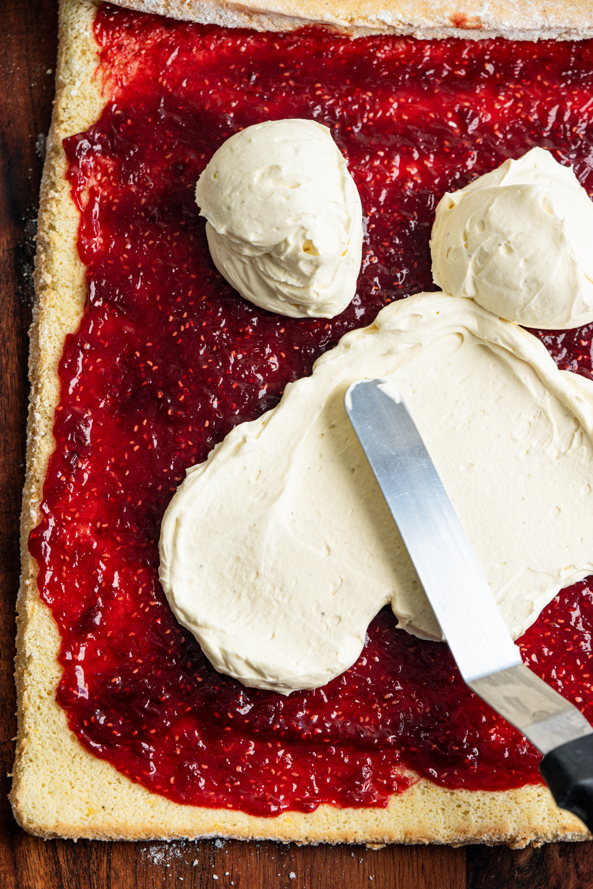 A spatula spreads cream over a layer of red jam on a rectangular sheet of sponge cake, preparing it for rolling.