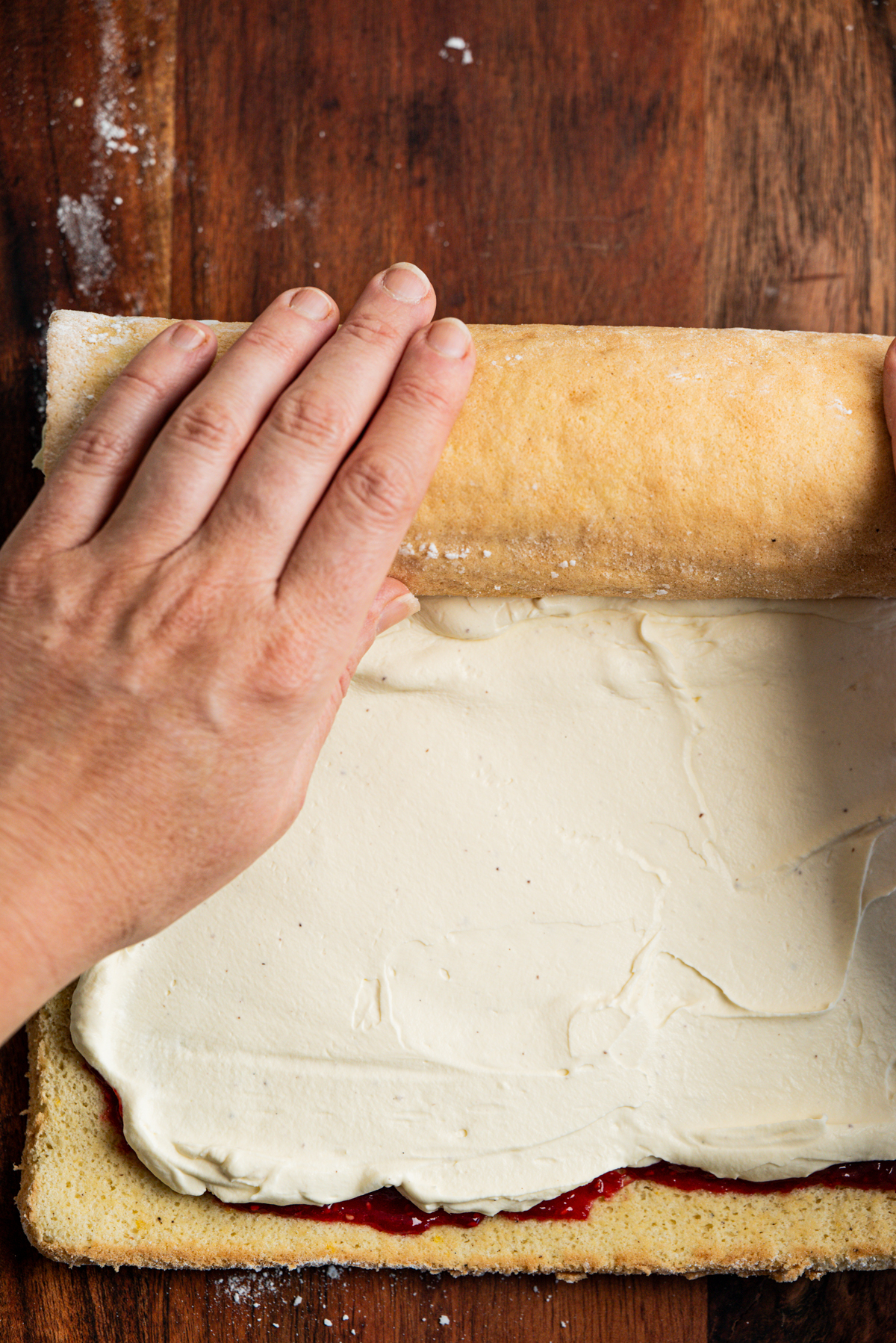 Two hands roll a sponge cake with a creamy white filling and a layer of red jam on a wooden surface.