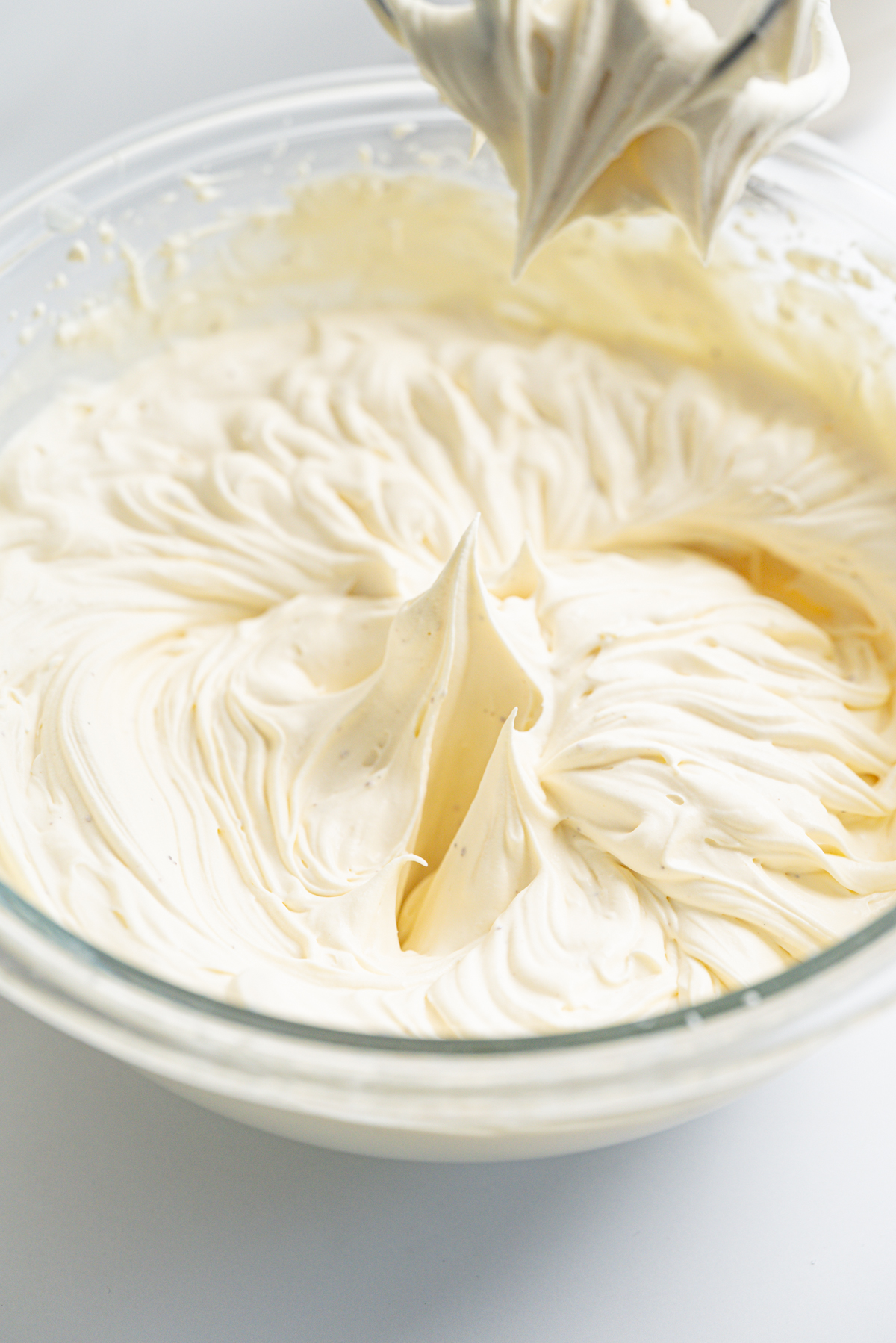 A glass bowl filled with smooth, creamy whipped mixture. Peaks have formed on the surface, indicating it has been freshly whipped. A mixer with more cream is held above the bowl.