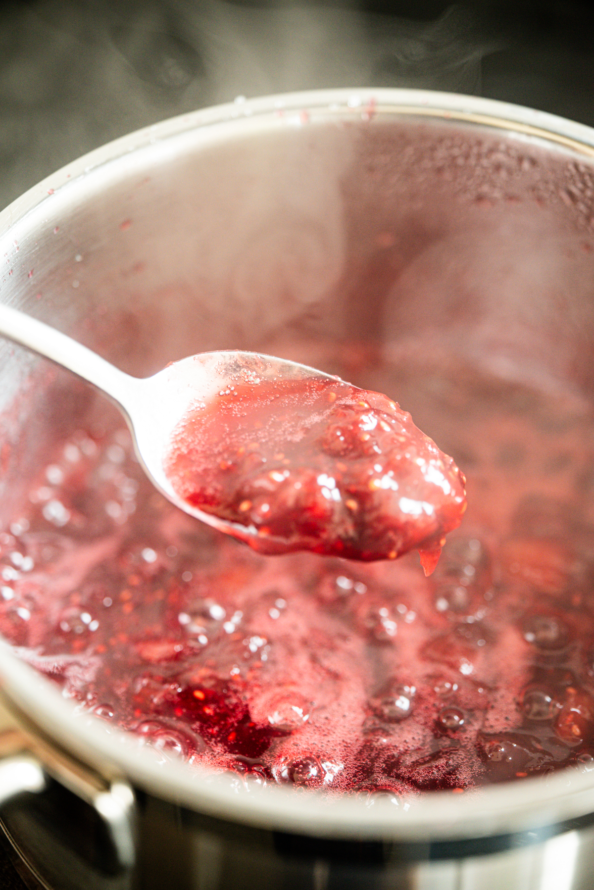 A spoon holds up a portion of bubbling, thick red cranberry sauce over a saucepan filled with more sauce, with steam rising from the pot.
