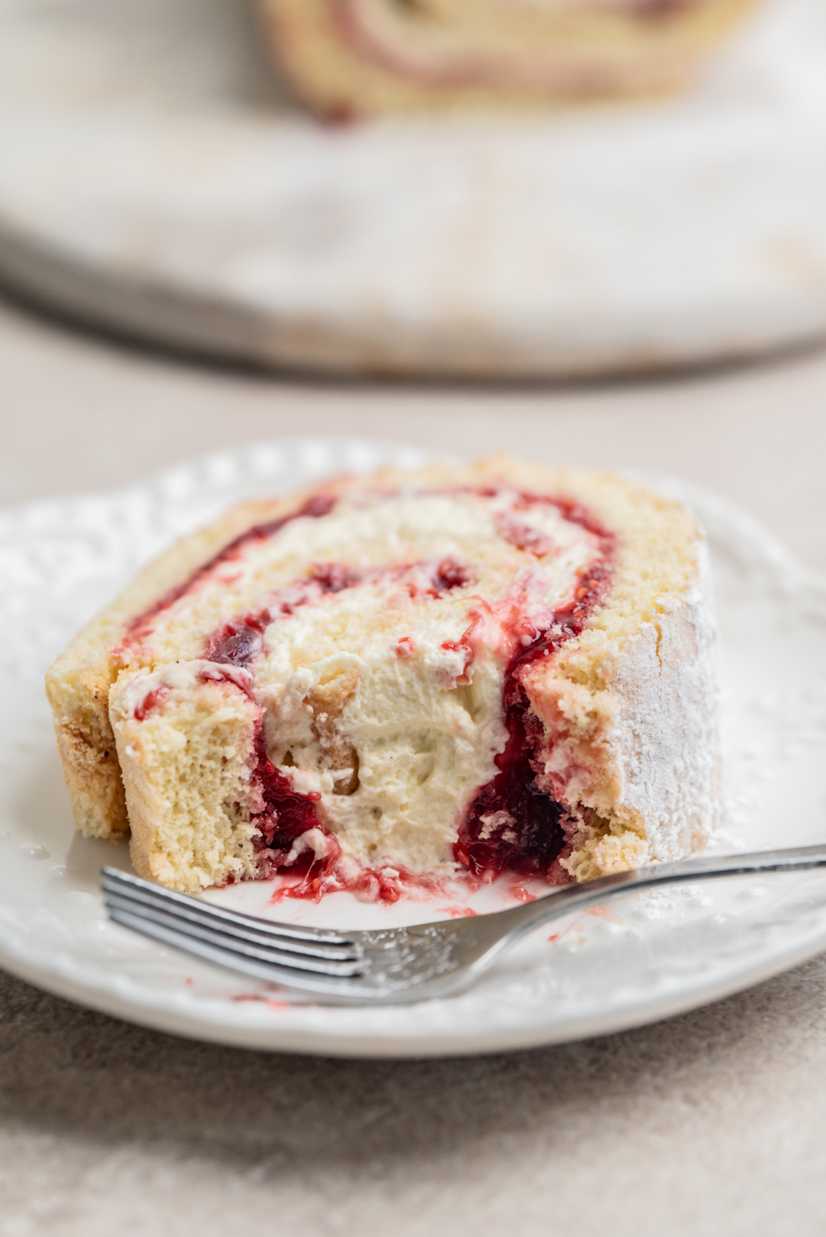 A slice of jelly roll cake filled with cream and rhubarb raspberry jam sits on a white plate, partially eaten, with a fork beside it.