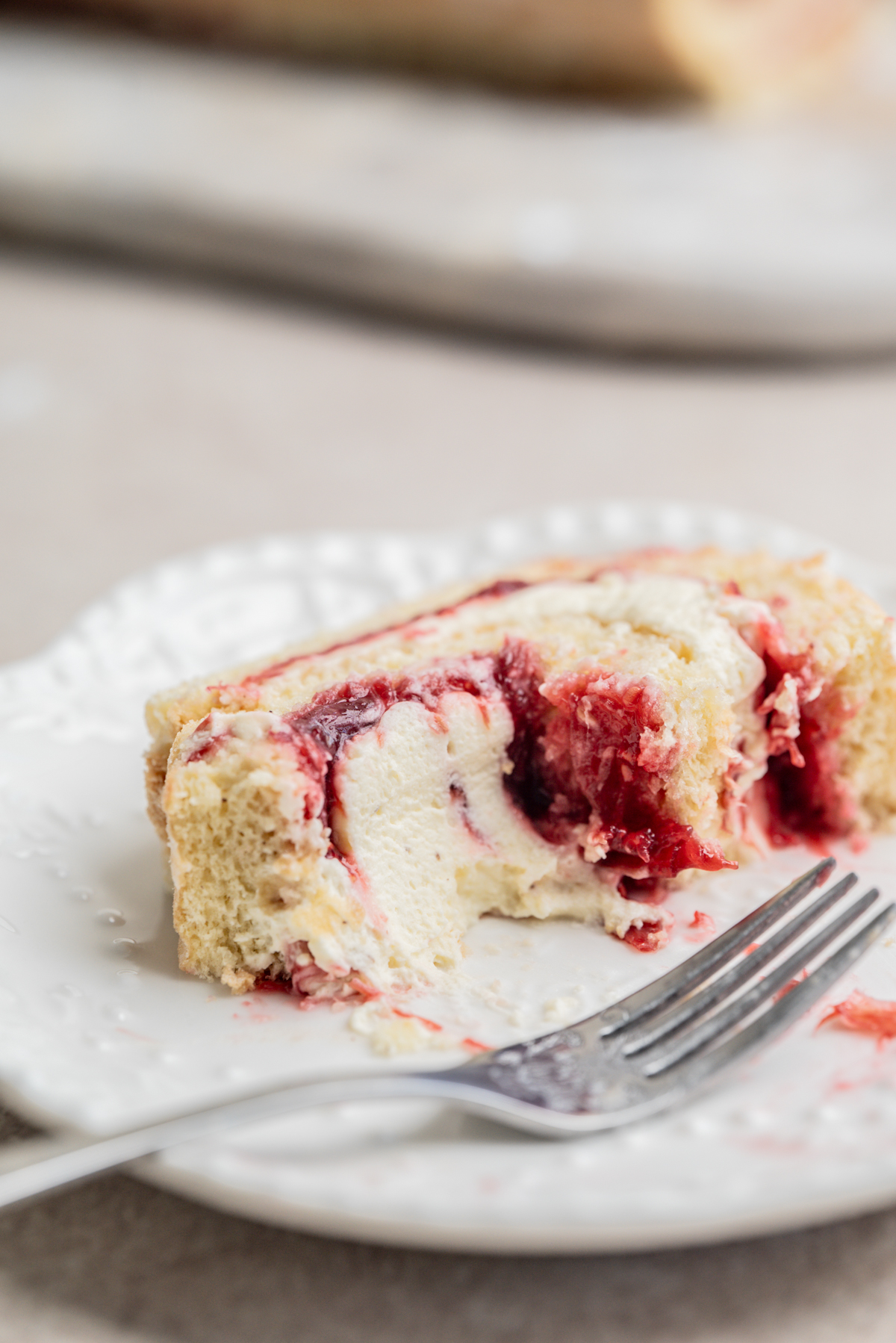 A slice of rolled cake with cream filling and raspberry or strawberry jam on a white plate, with a fork placed in front.
