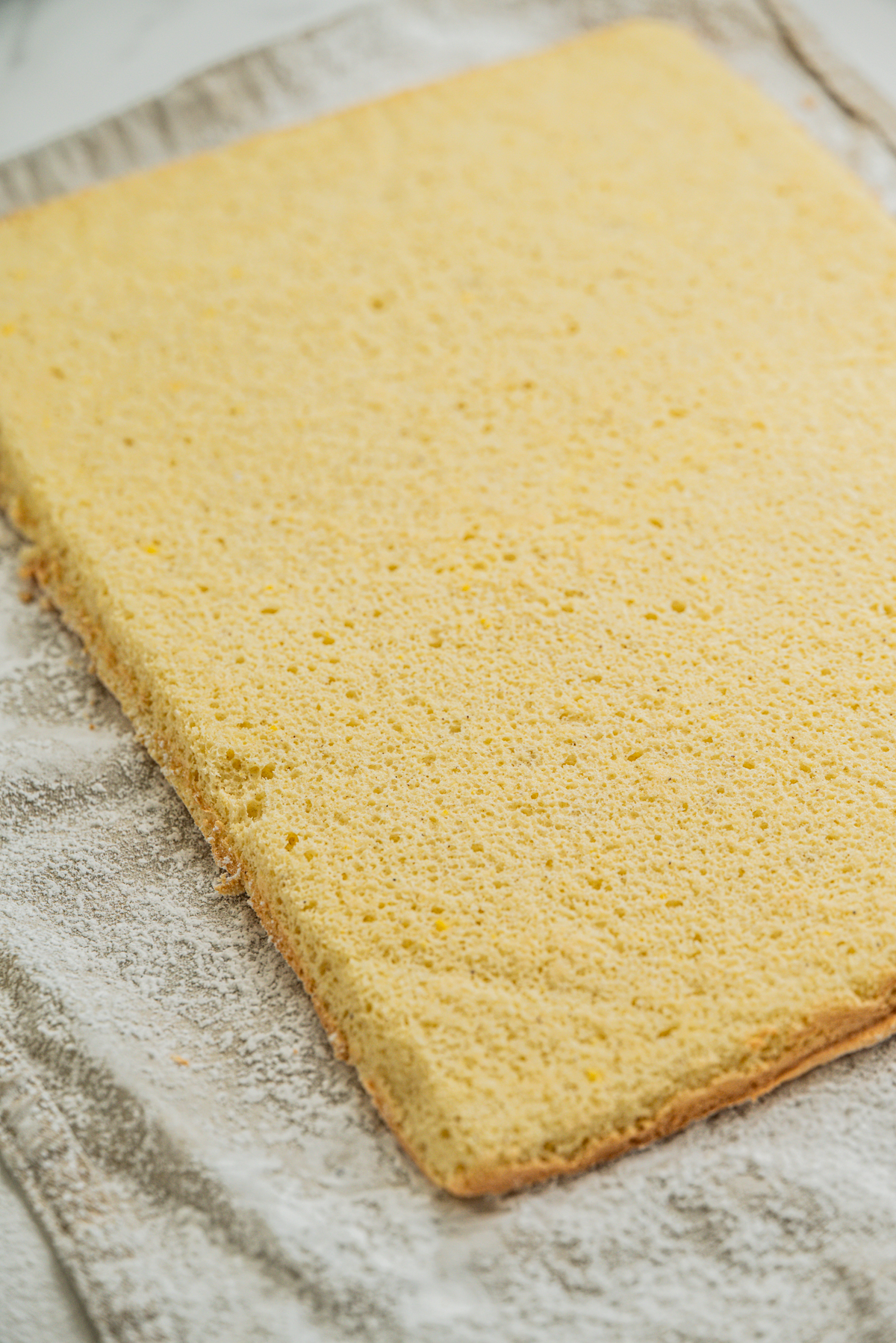 A rectangular sheet of plain yellow sponge cake rests on a light-colored, floured kitchen towel.