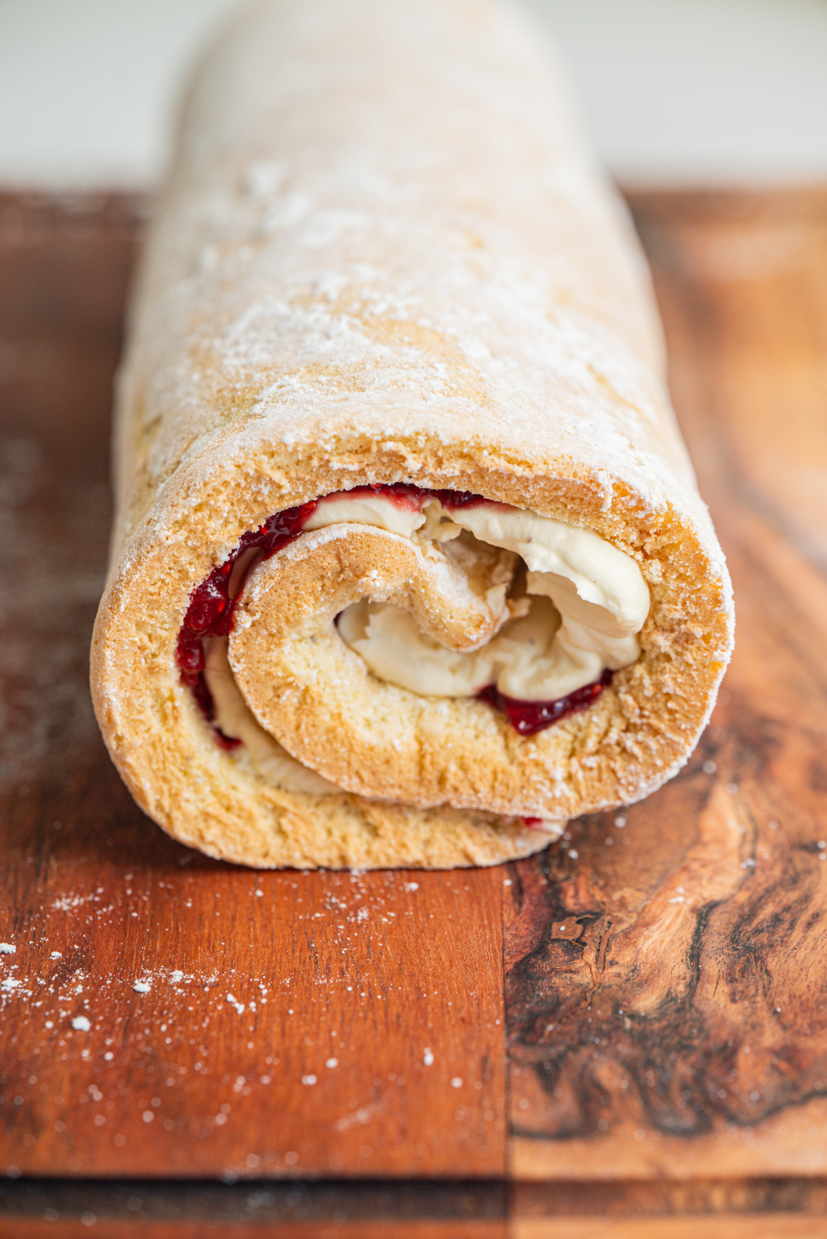 A close-up of a jelly roll cake filled with cream and rhubarb jam, resting on a wooden cutting board, lightly dusted with powdered sugar.