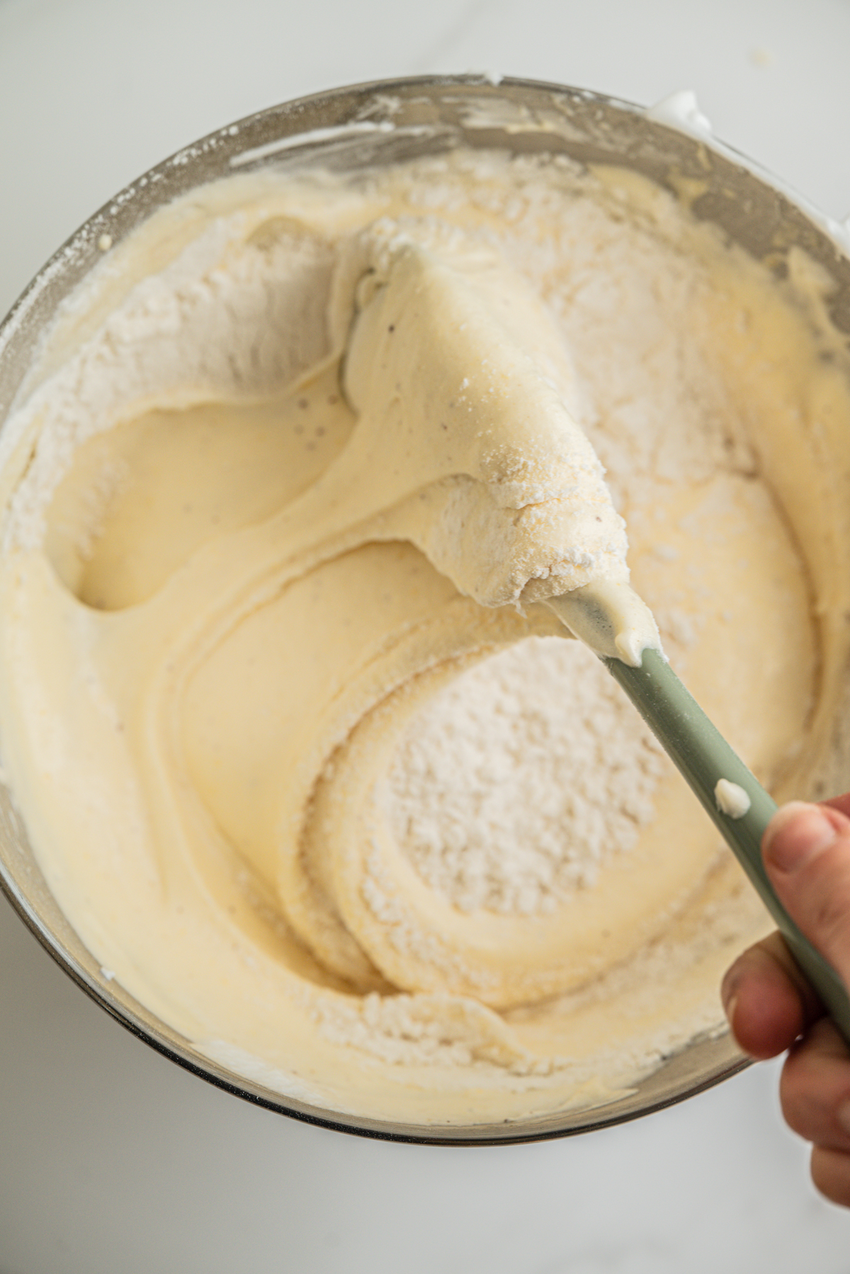 A hand uses a green spatula to fold flour into a pale yellow cake batter in a metal mixing bowl.
