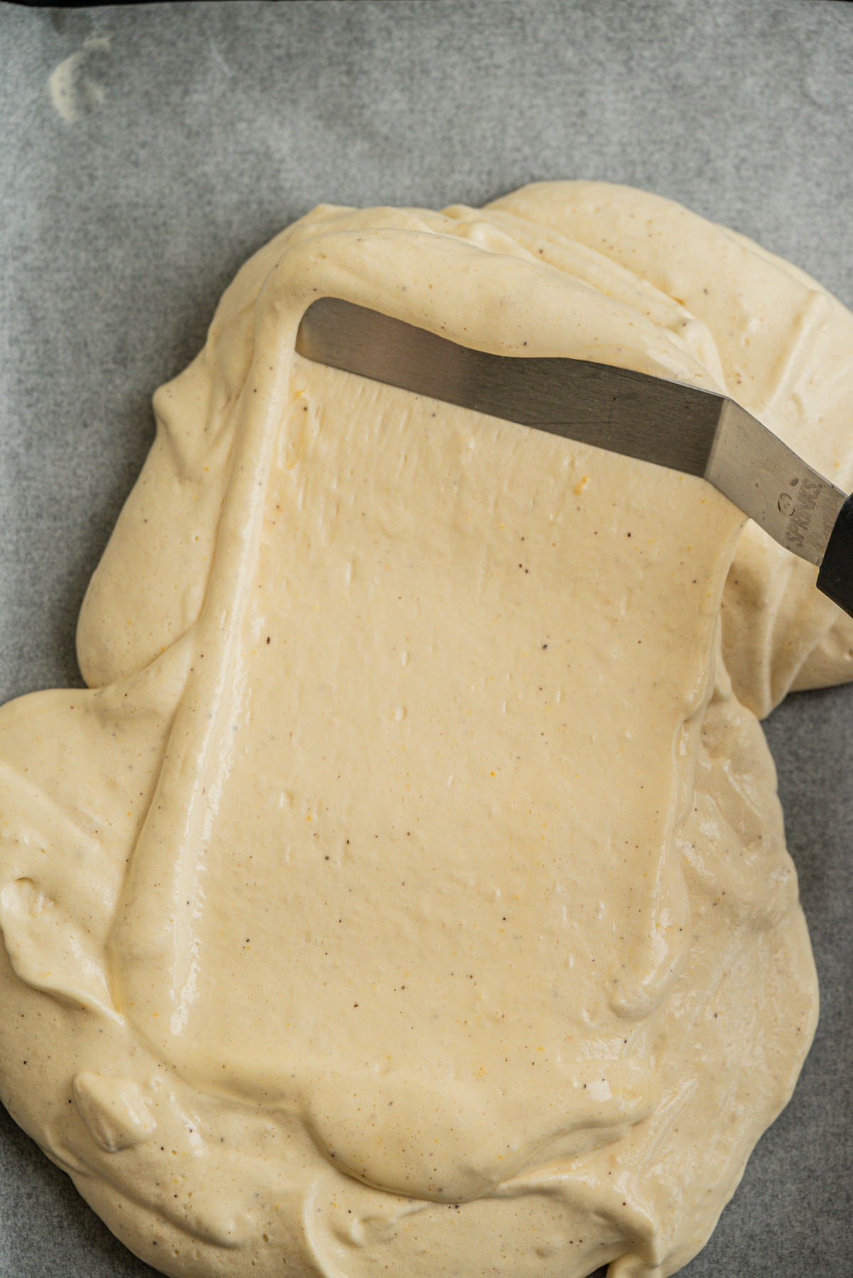 Batter being spread with a spatula on parchment paper, likely for baking a cake or dessert.