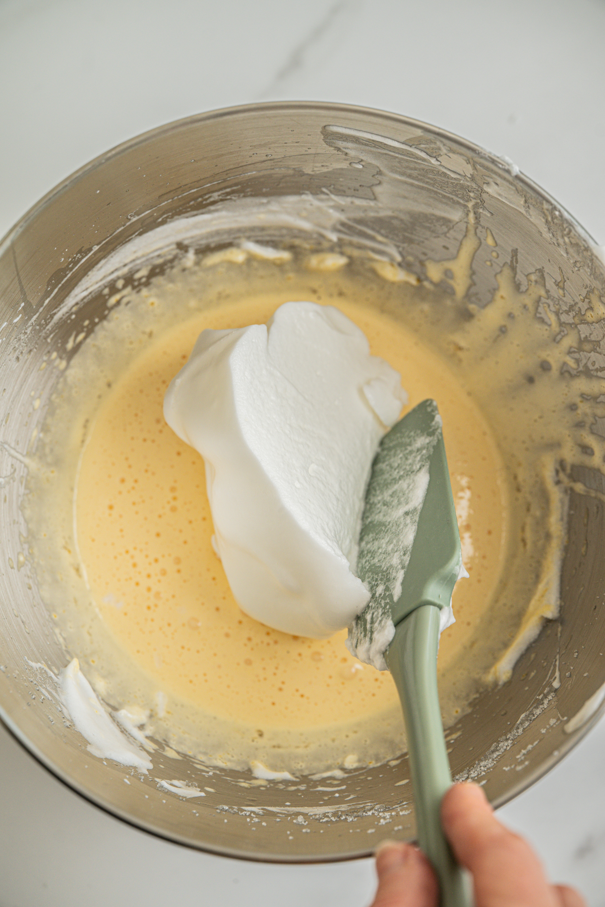 A hand uses a green spatula to fold fluffy whipped egg whites into a yellow batter in a metal mixing bowl on a white countertop.