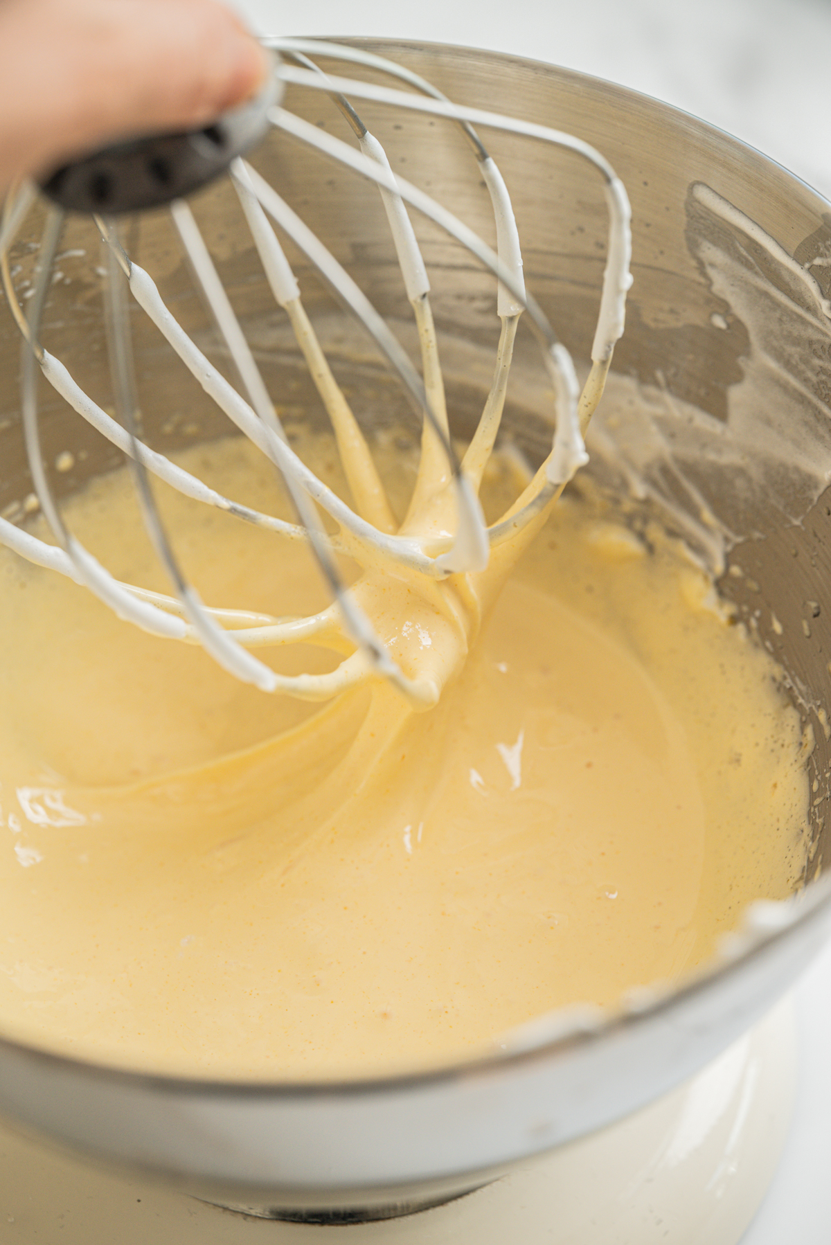 A close-up of a mixing bowl with thick, creamy batter being mixed by a wire whisk attachment, held above the bowl with some batter dripping off.