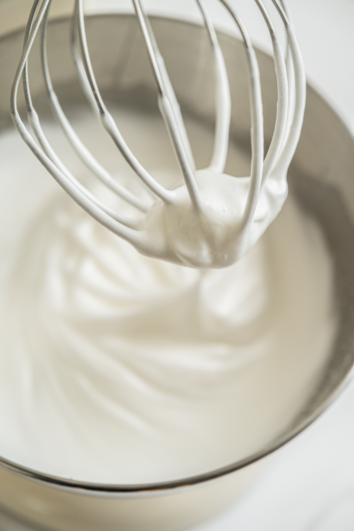 A metal whisk attachment with glossy, stiff peaks of whipped egg whites in a stainless steel mixing bowl.