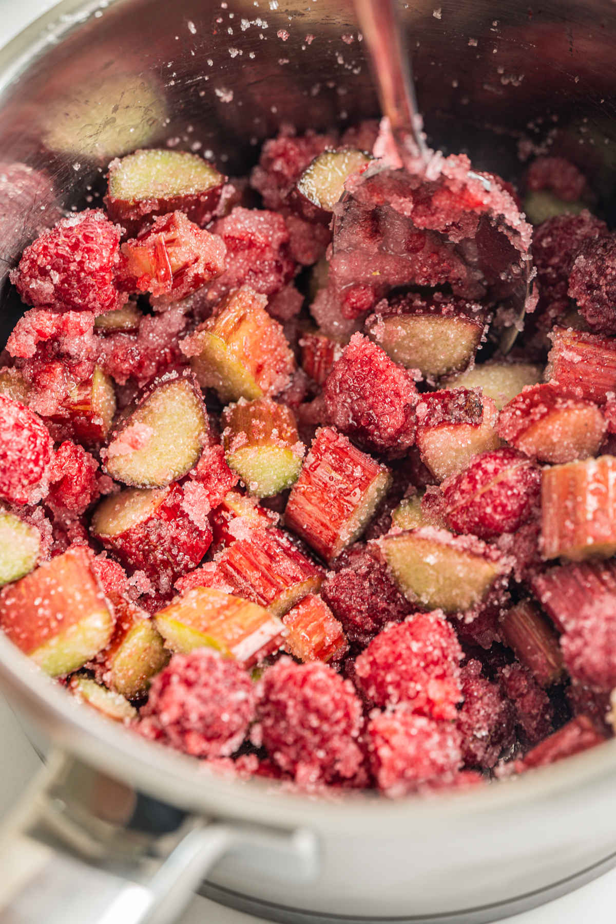 Chopped rhubarb and strawberries mixed with sugar in a metal saucepan, with a spoon partially visible, ready to be cooked for a dessert or jam.
