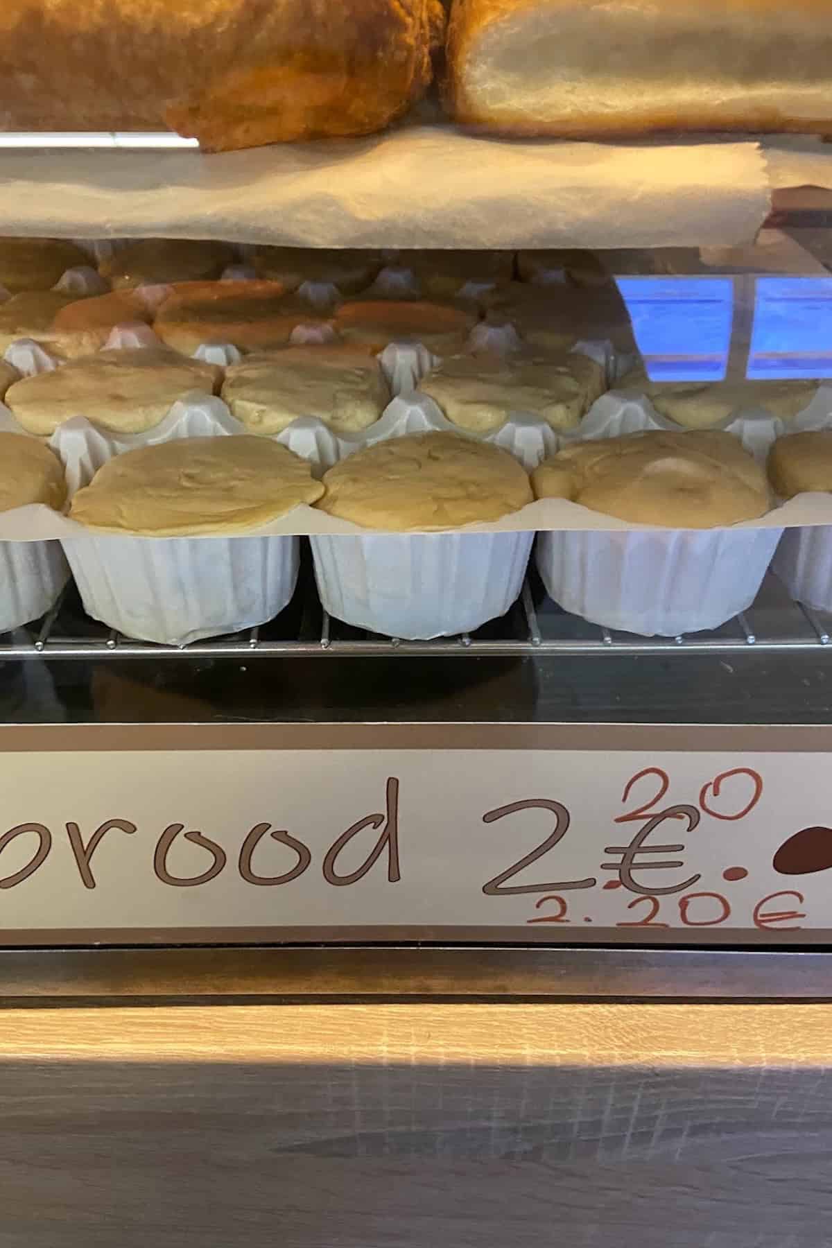 Several round bread rolls in white paper cups are displayed on a bakery shelf, with a sign below reading 2.20€ (crossed out) and 2€ for the price.