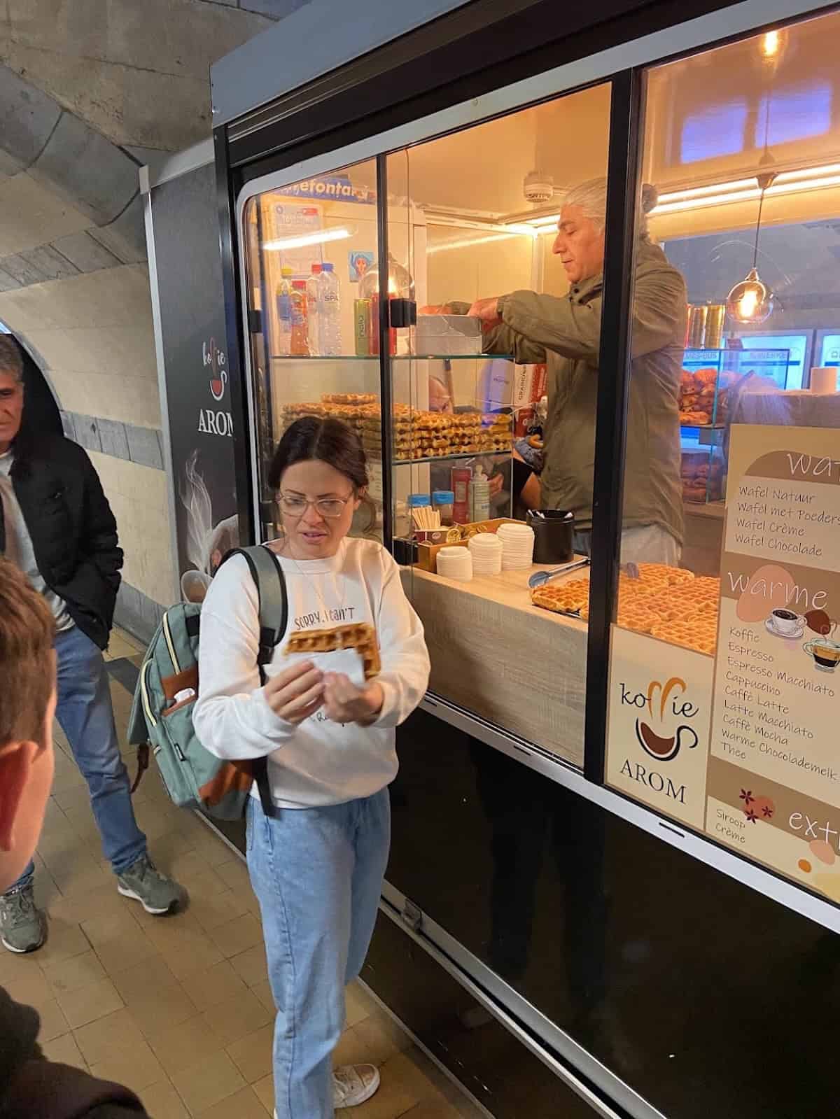 A woman holding a treat stands near a coffee and snack kiosk, while a vendor serves customers inside the kiosk. Other people wait nearby.