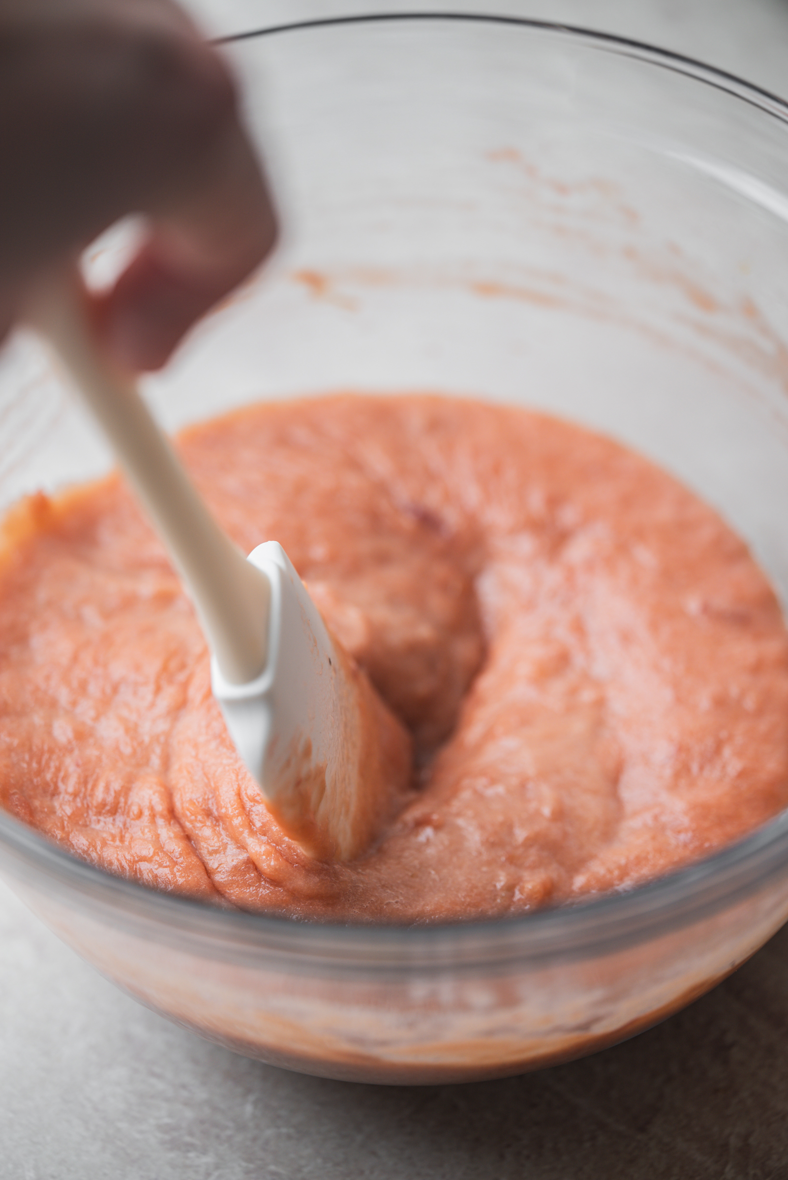 A hand stirs a thick rhubarb curd in a clear glass bowl using a white spatula.