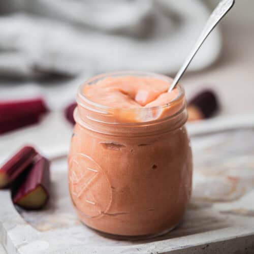 A glass jar filled with smooth, pink rhubarb curd sits on a marble board. A spoon is in the jar, and chopped rhubarb pieces are scattered around. The background is softly blurred.