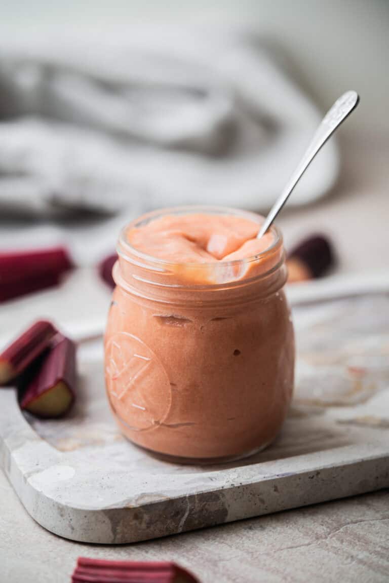 A glass jar filled with smooth, pink rhubarb curd sits on a marble board. A spoon is in the jar, and chopped rhubarb pieces are scattered around. The background is softly blurred.