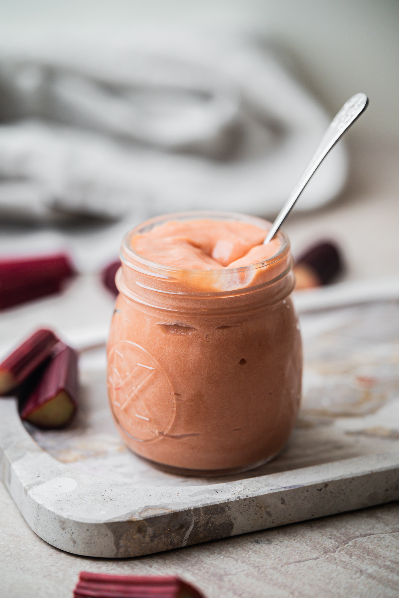 A glass jar filled with smooth, pink rhubarb curd sits on a marble board. A spoon is in the jar, and chopped rhubarb pieces are scattered around. The background is softly blurred.