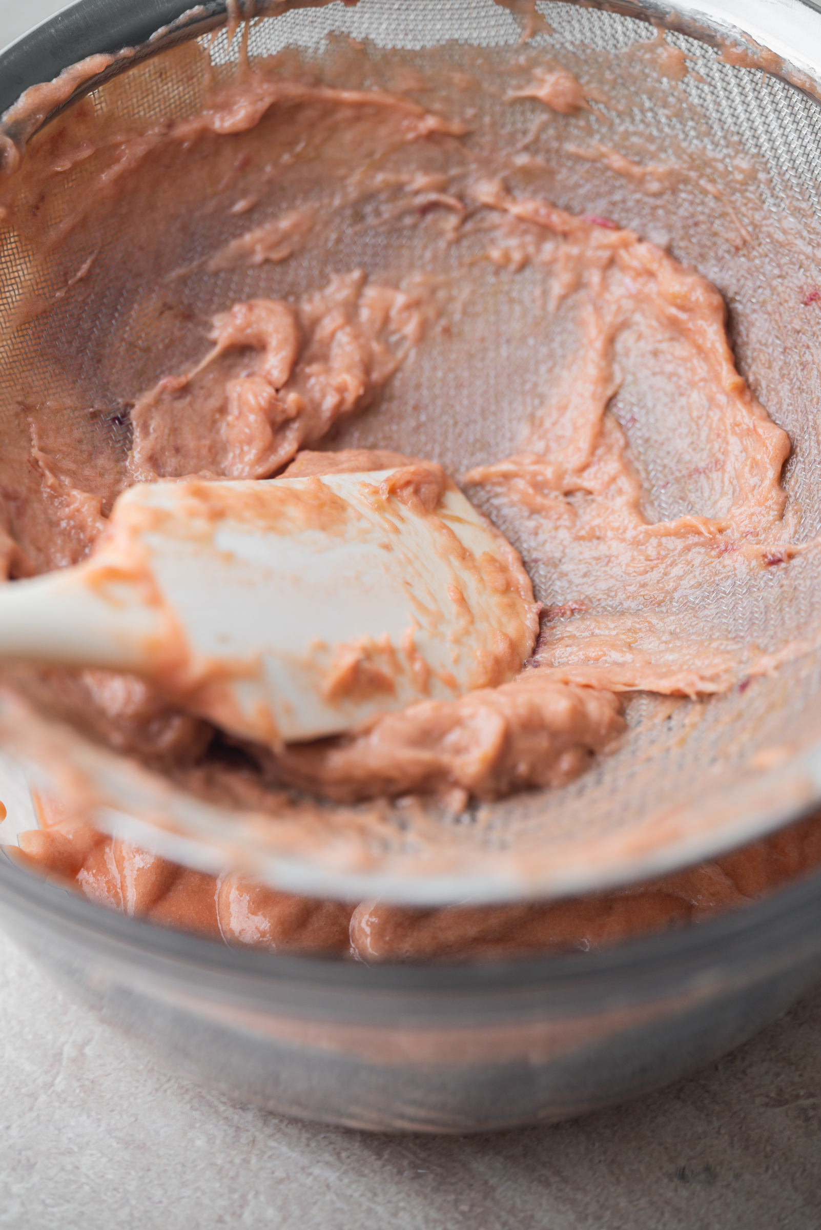 A metal bowl filled with light pink rhubarb curd being mixed or scraped with a white rubber spatula, sits on a light-colored surface.