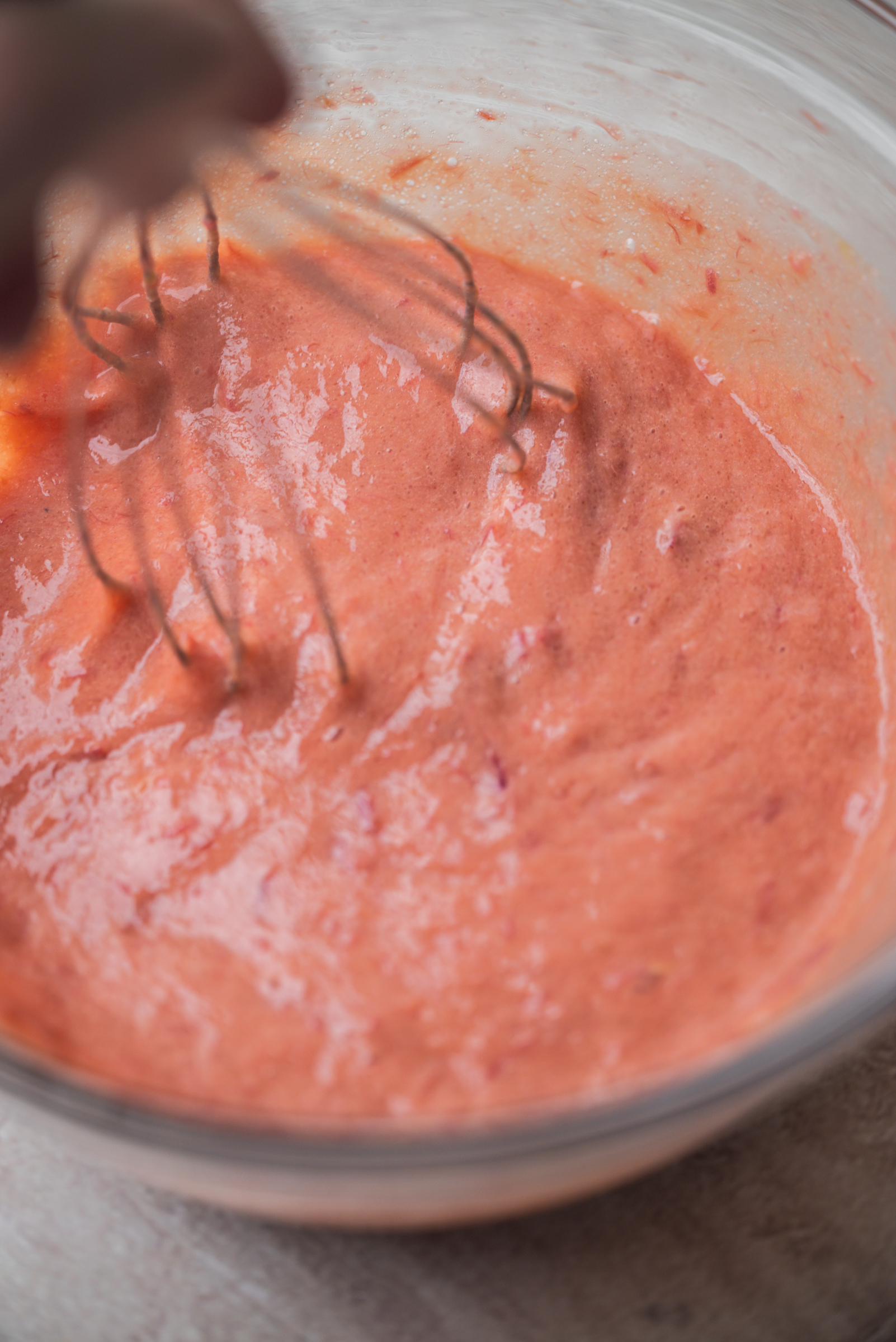 A close-up of a hand whisking pink rhubarb curd in a clear glass bowl on a light-colored surface.