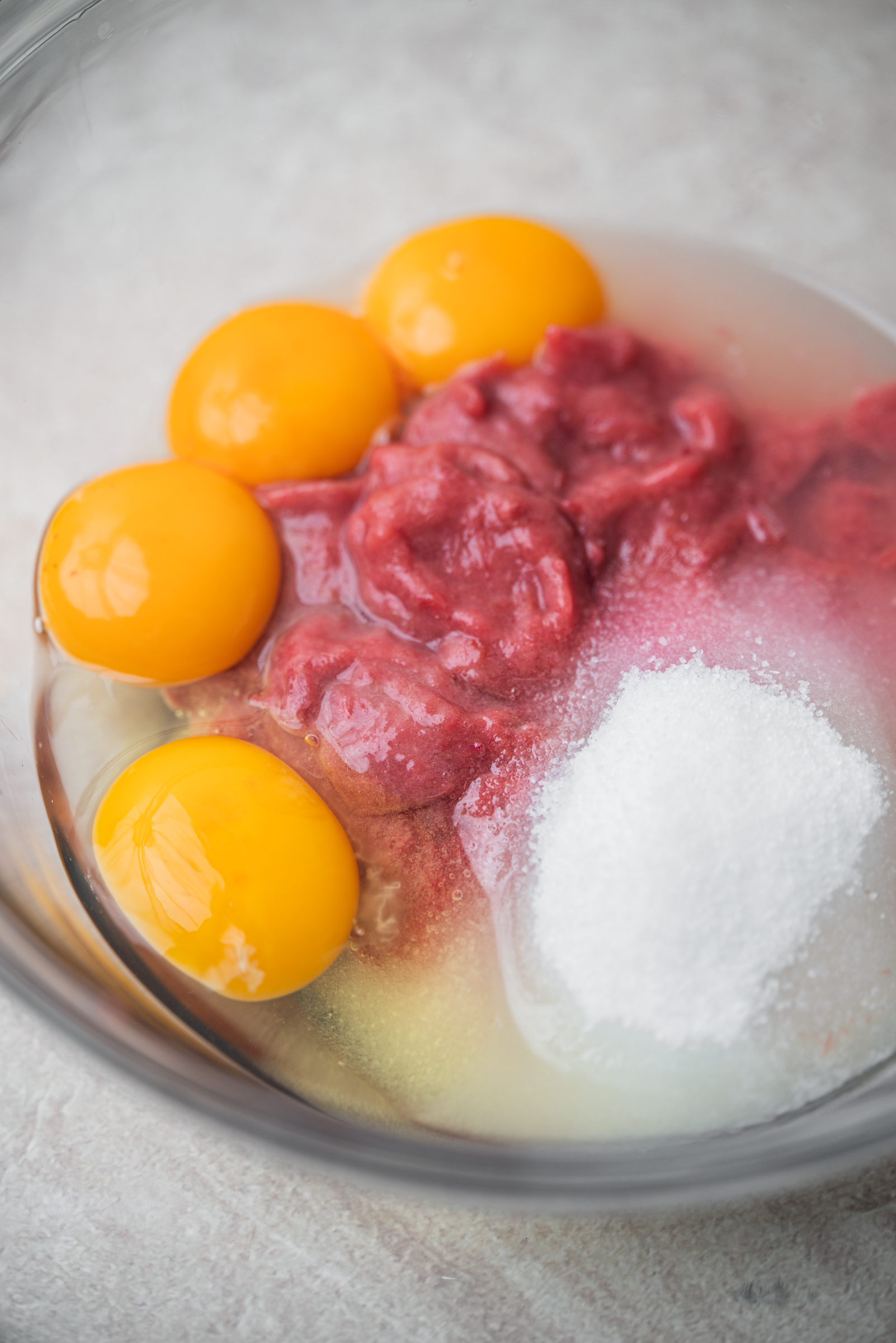 Four egg yolks, rhubarb compote, and a mound of white sugar are in a glass bowl, ready to be mixed, all sitting on a light-colored surface.