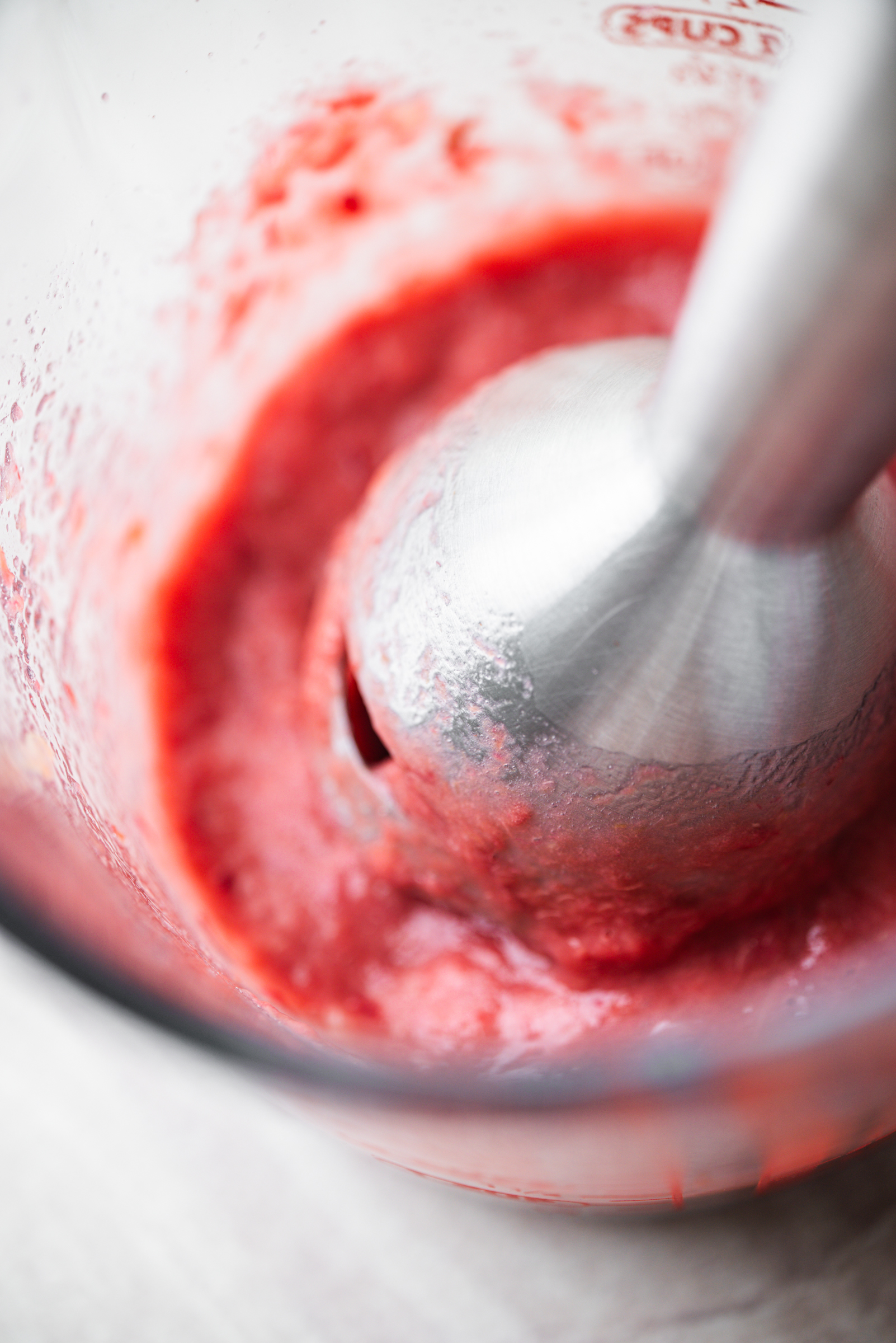 A close-up of a hand blender pureeing a bright red rhubarb inside a clear glass container.