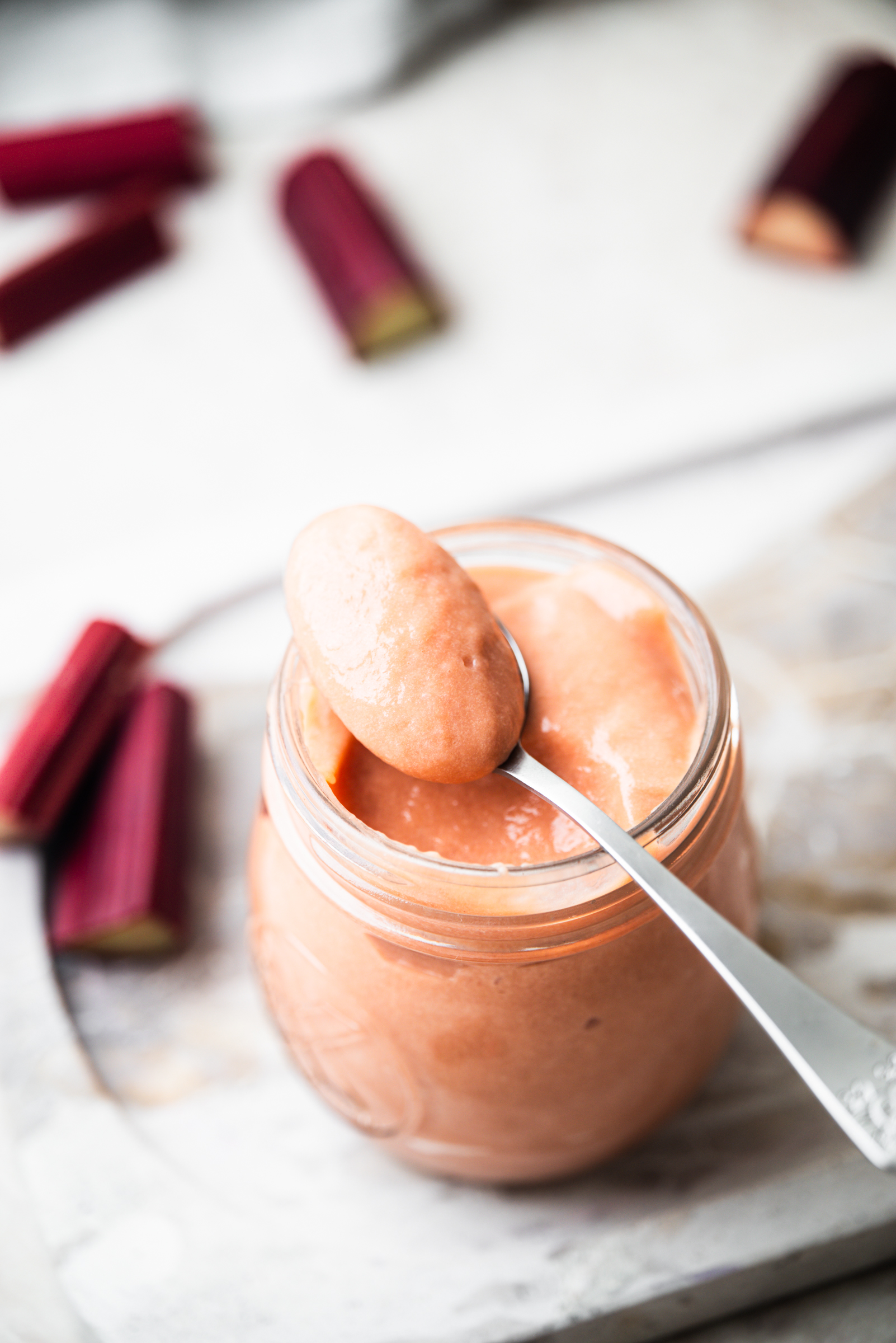 A spoonful of pink rhubarb sauce resting over a glass jar filled with the same sauce, with pieces of raw rhubarb in the background on a white surface.