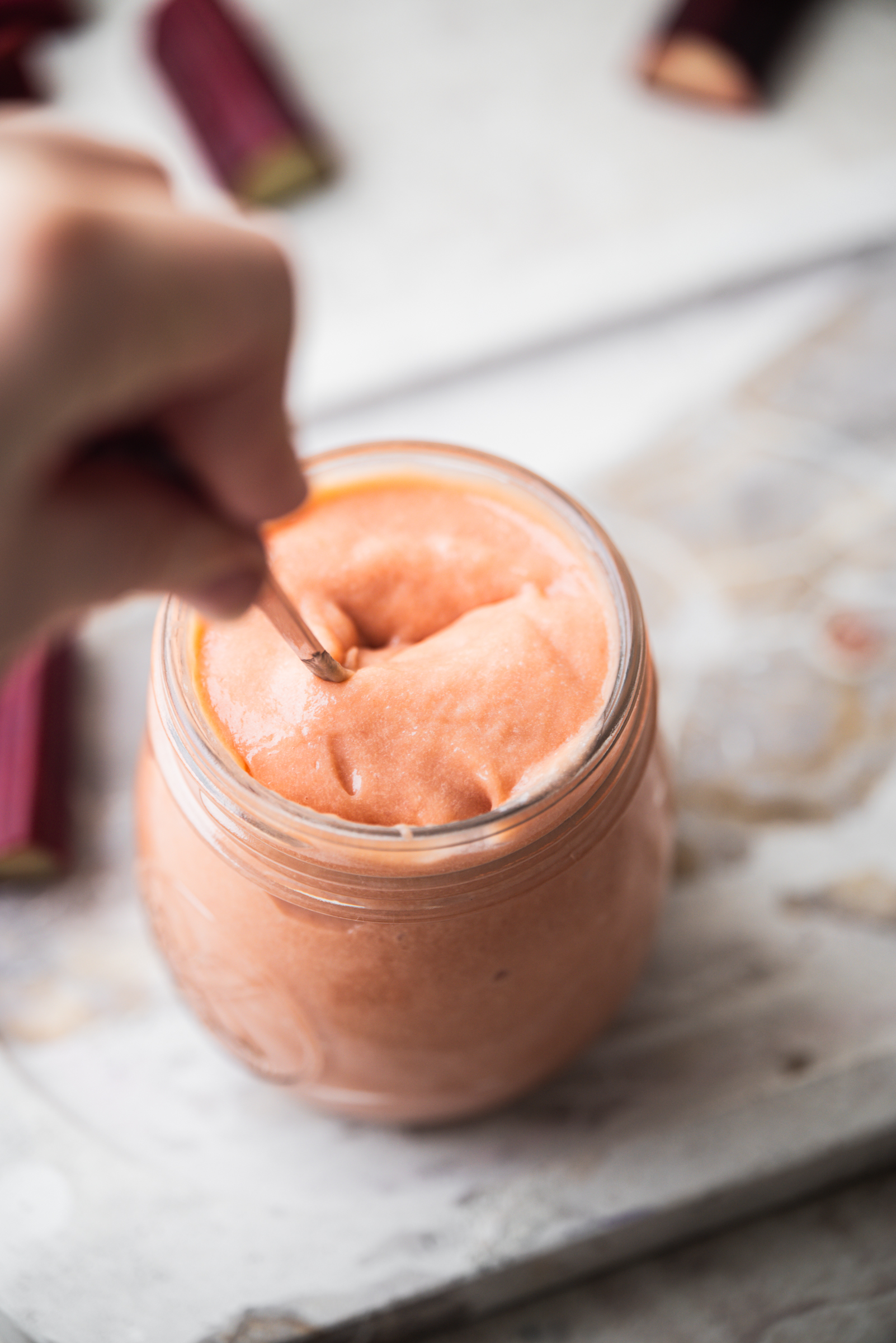 A hand stirs creamy, light rhubarb curd in a glass jar. Blurred pieces of rhubarb are visible in the background on a light-colored surface.