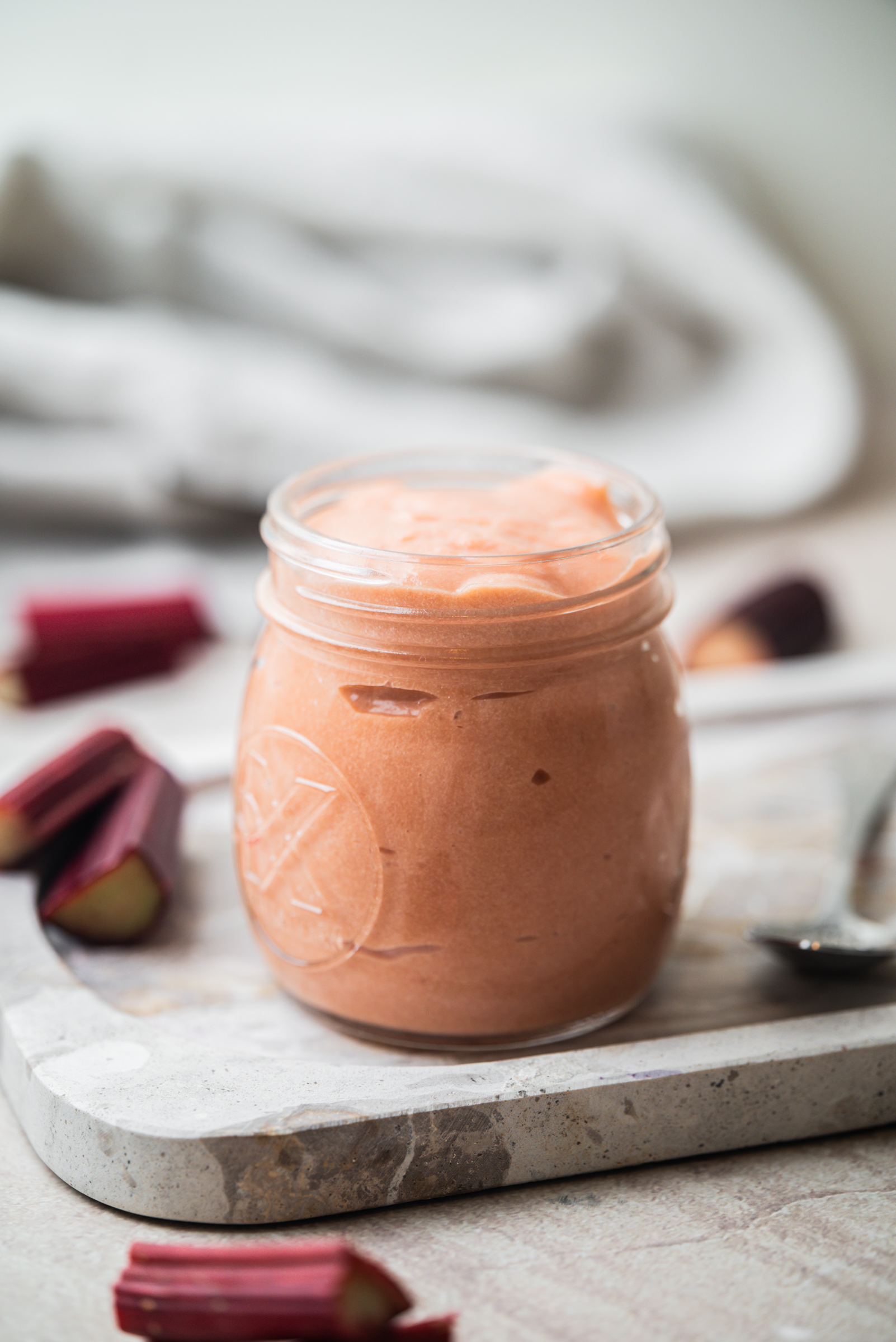 A small glass jar filled with creamy, light pink rhubarb curd sits on a marble board, with fresh rhubarb pieces and a spoon nearby.