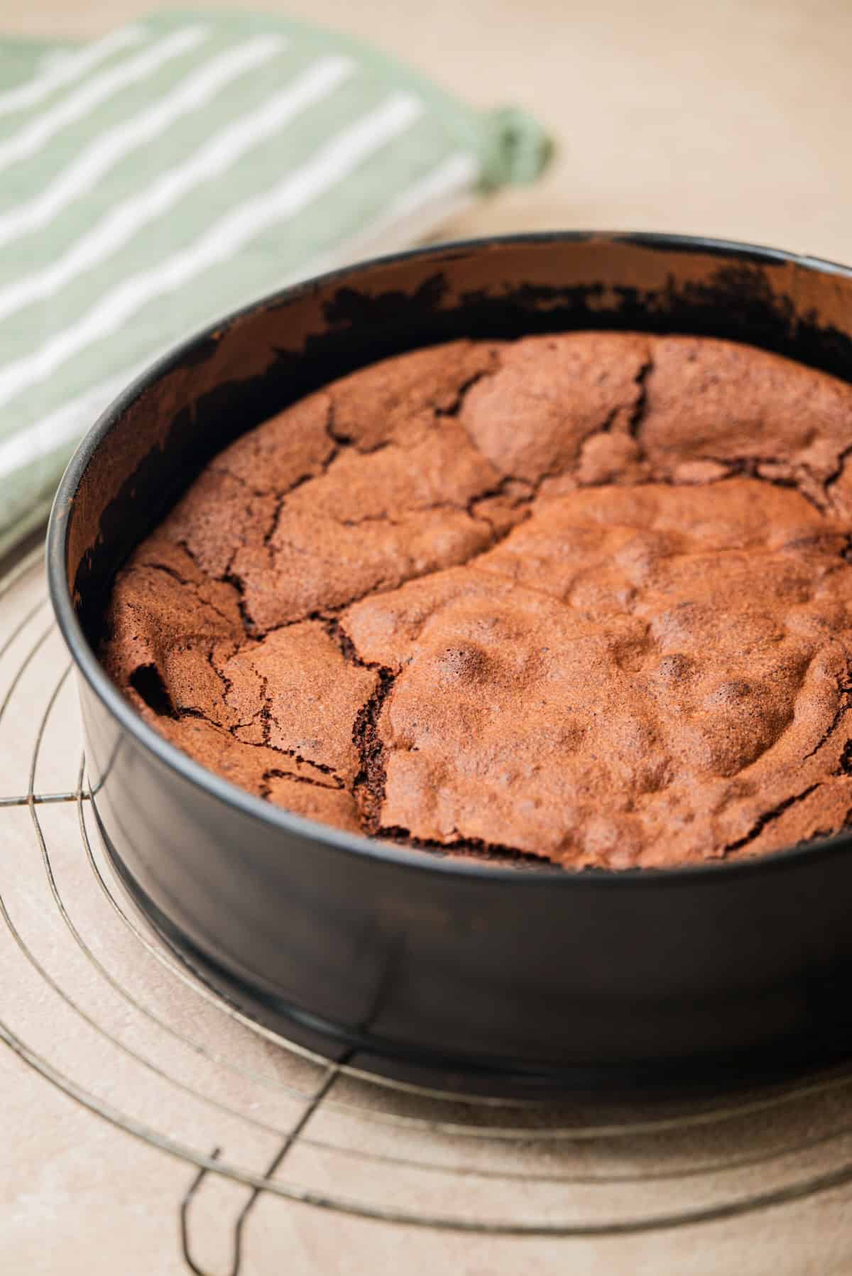 A freshly baked chocolate cake with a cracked top sits in a round black baking pan on a wire rack, next to a green and white striped kitchen towel.
