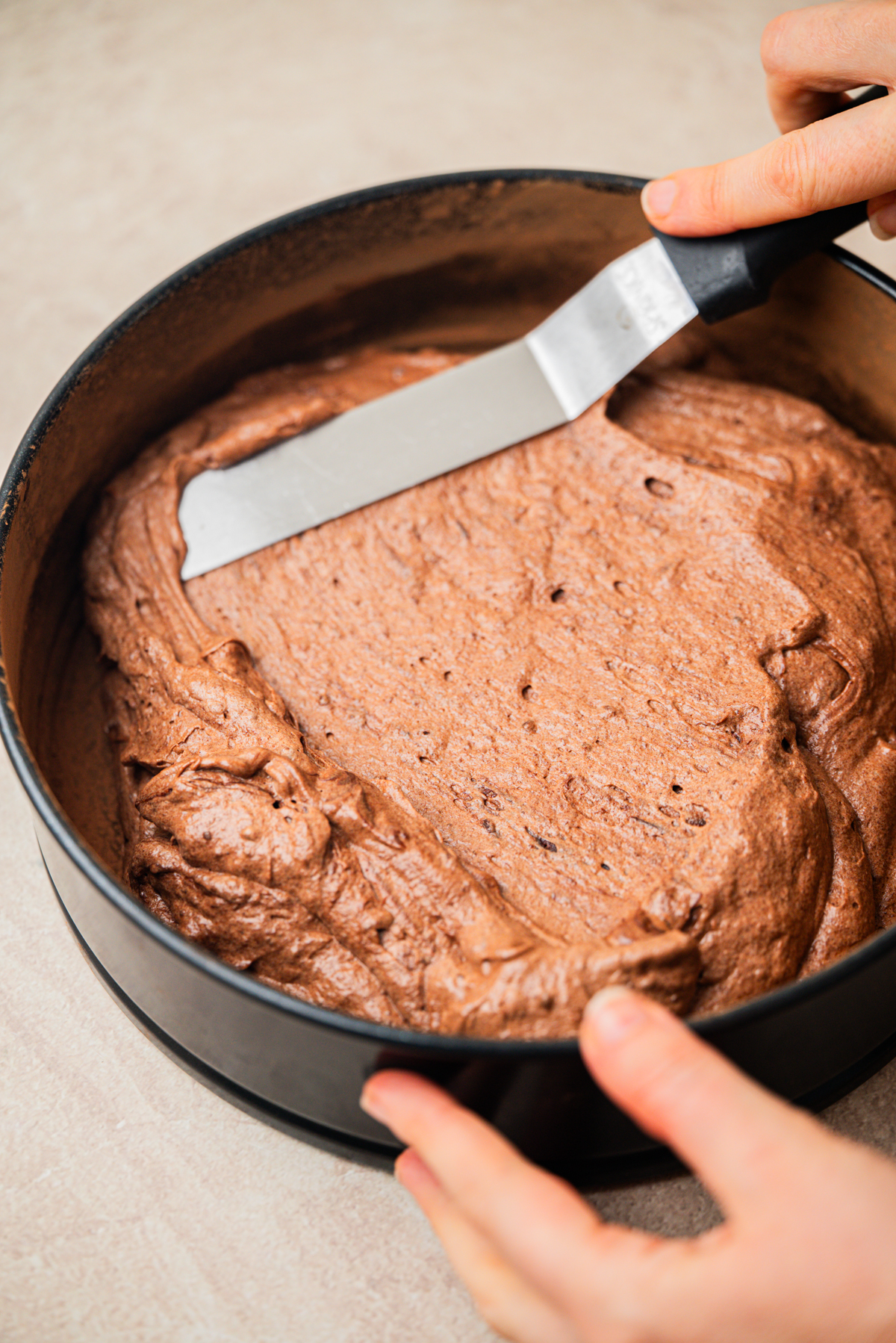 Hands spreading chocolate cake batter in a round baking pan with an offset spatula on a light-colored surface.