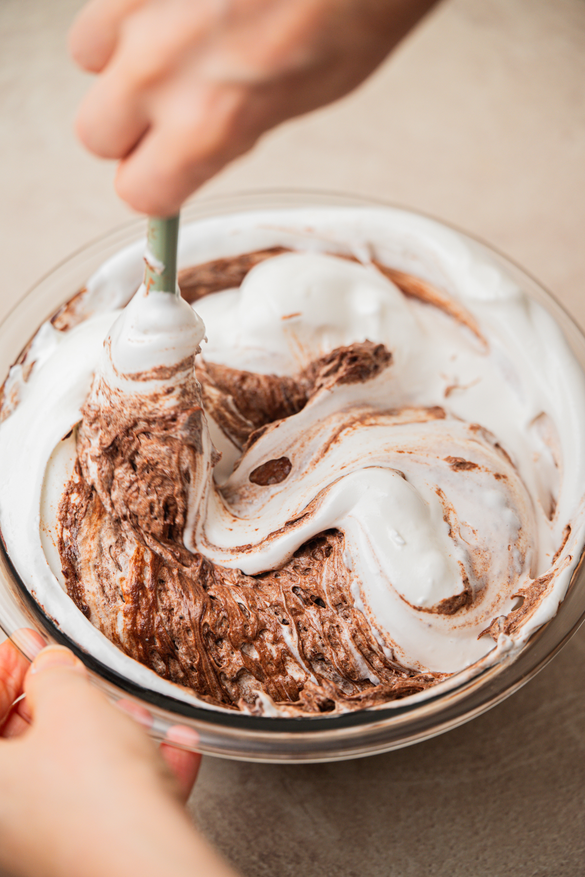 Hands mix fluffy white whipped cream and chocolate in a glass bowl with a spatula, creating a marbled effect. The bowl is placed on a light-colored surface.