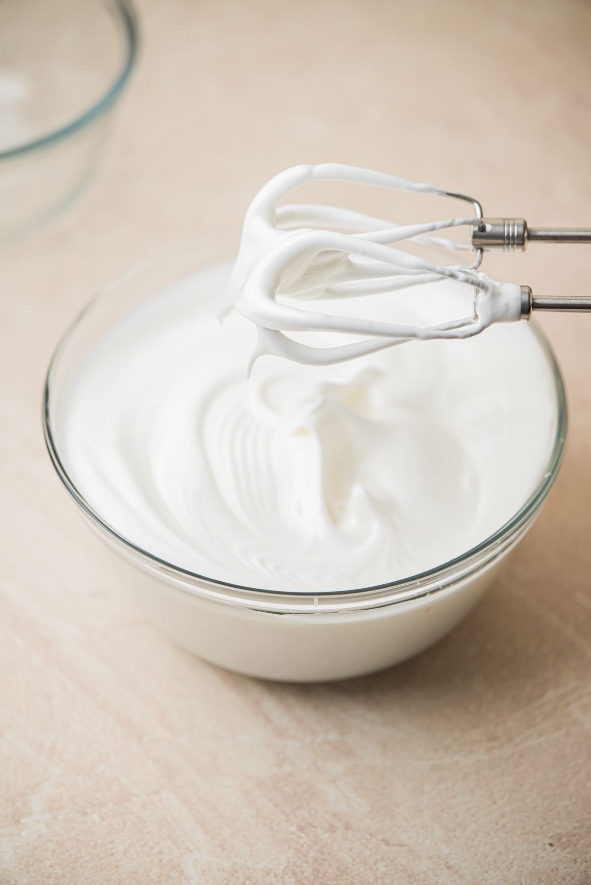 A glass bowl filled with fluffy, white whipped cream, with a hand mixer beater covered in the whipped cream held above the bowl.