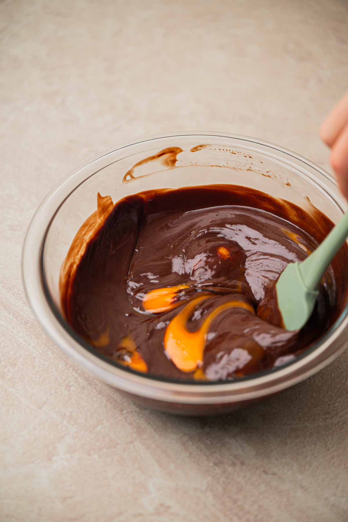 A hand stirs glossy, melted chocolate and butter with visible egg yolks in a clear glass bowl using a green spatula, on a light countertop.