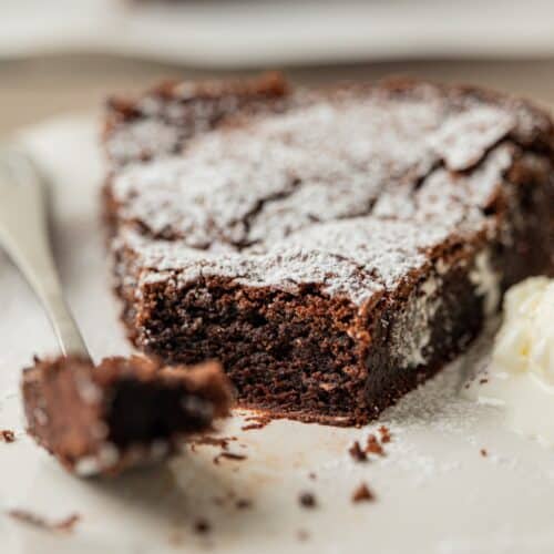 A close-up of a slice of chocolate cake topped with powdered sugar on a white plate, with a fork holding a small bite next to it.