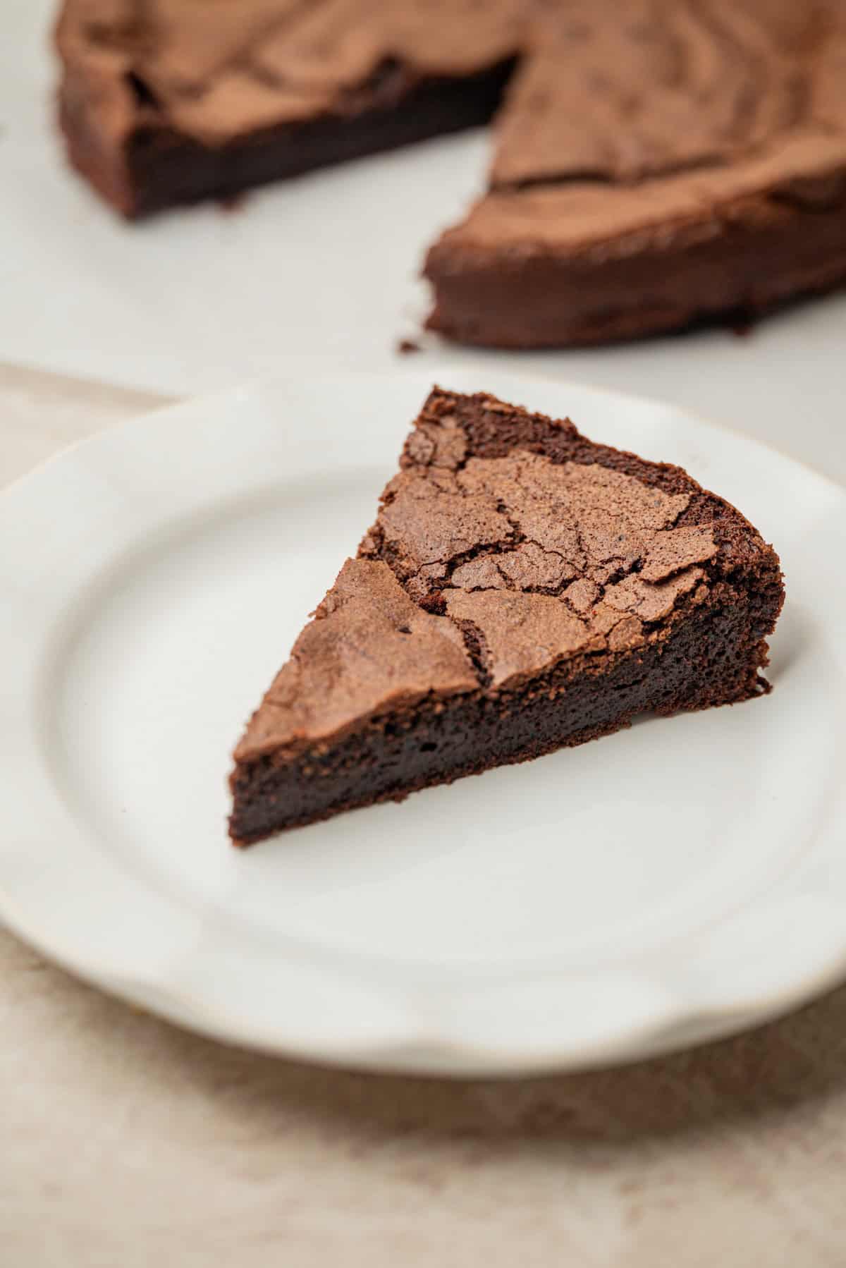 A slice of rich, dense chocolate cake with a crackled top sits on a white plate. The rest of the cake is visible in the background.
