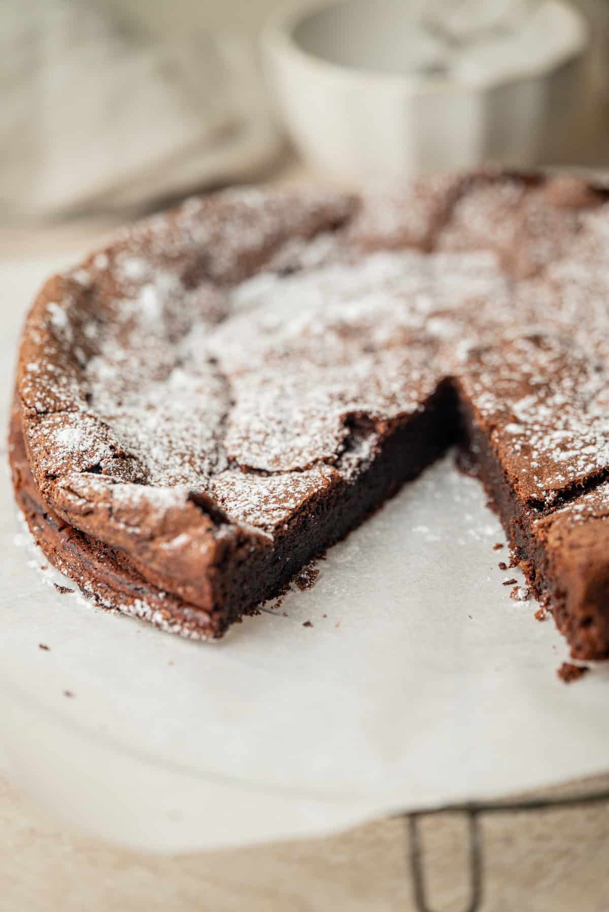 A round chocolate cake dusted with powdered sugar sits on parchment paper, with one slice missing. A white bowl is blurred in the background.