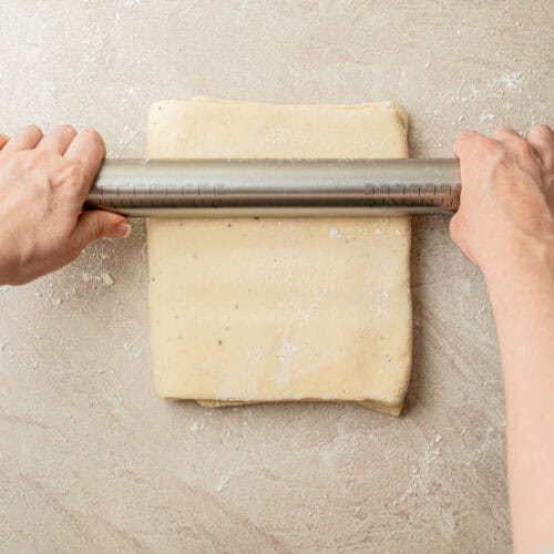 Hands use a metal rolling pin to flatten a square slab of dough on a lightly floured surface.