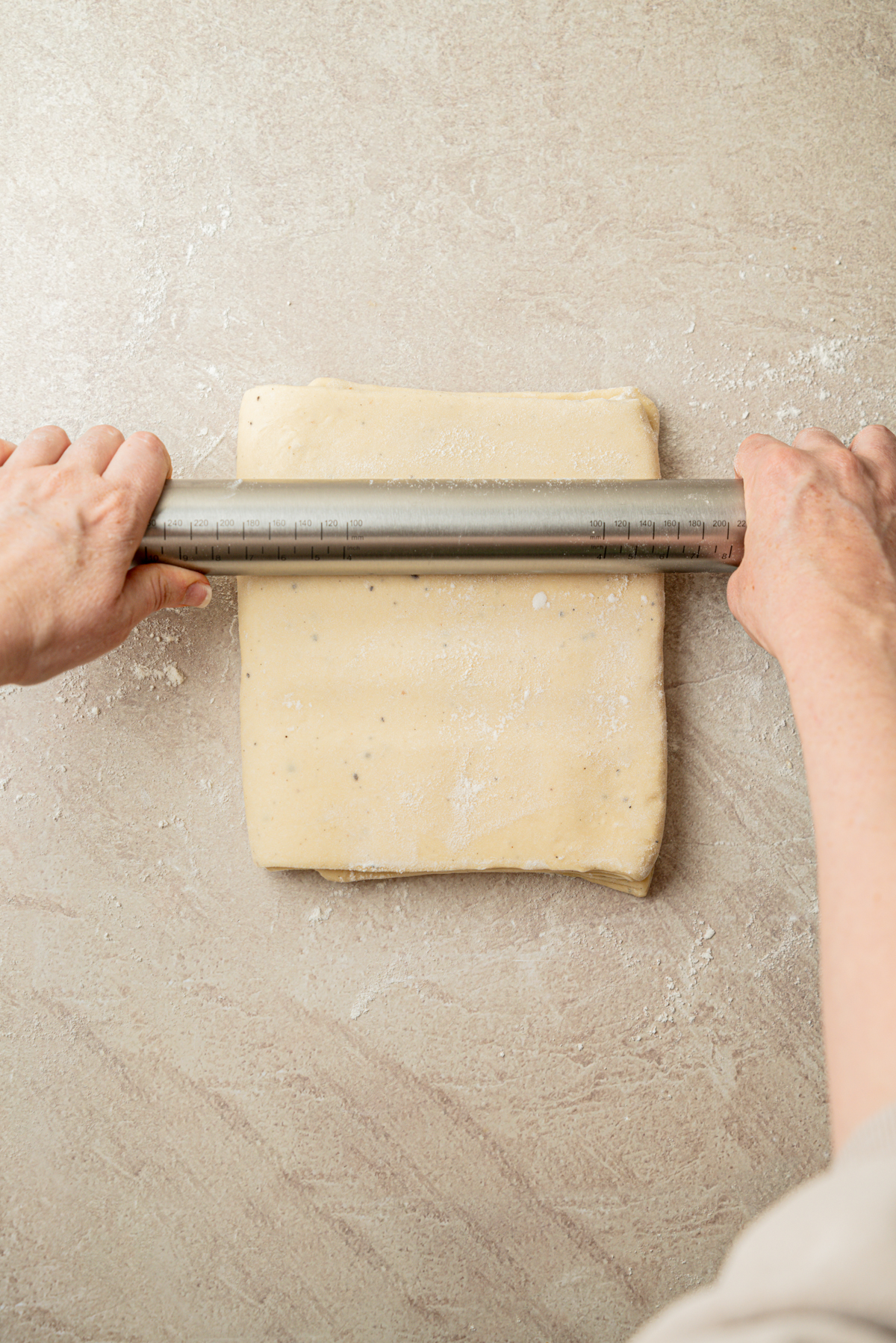 Hands use a metal rolling pin to flatten a square slab of dough on a lightly floured surface.